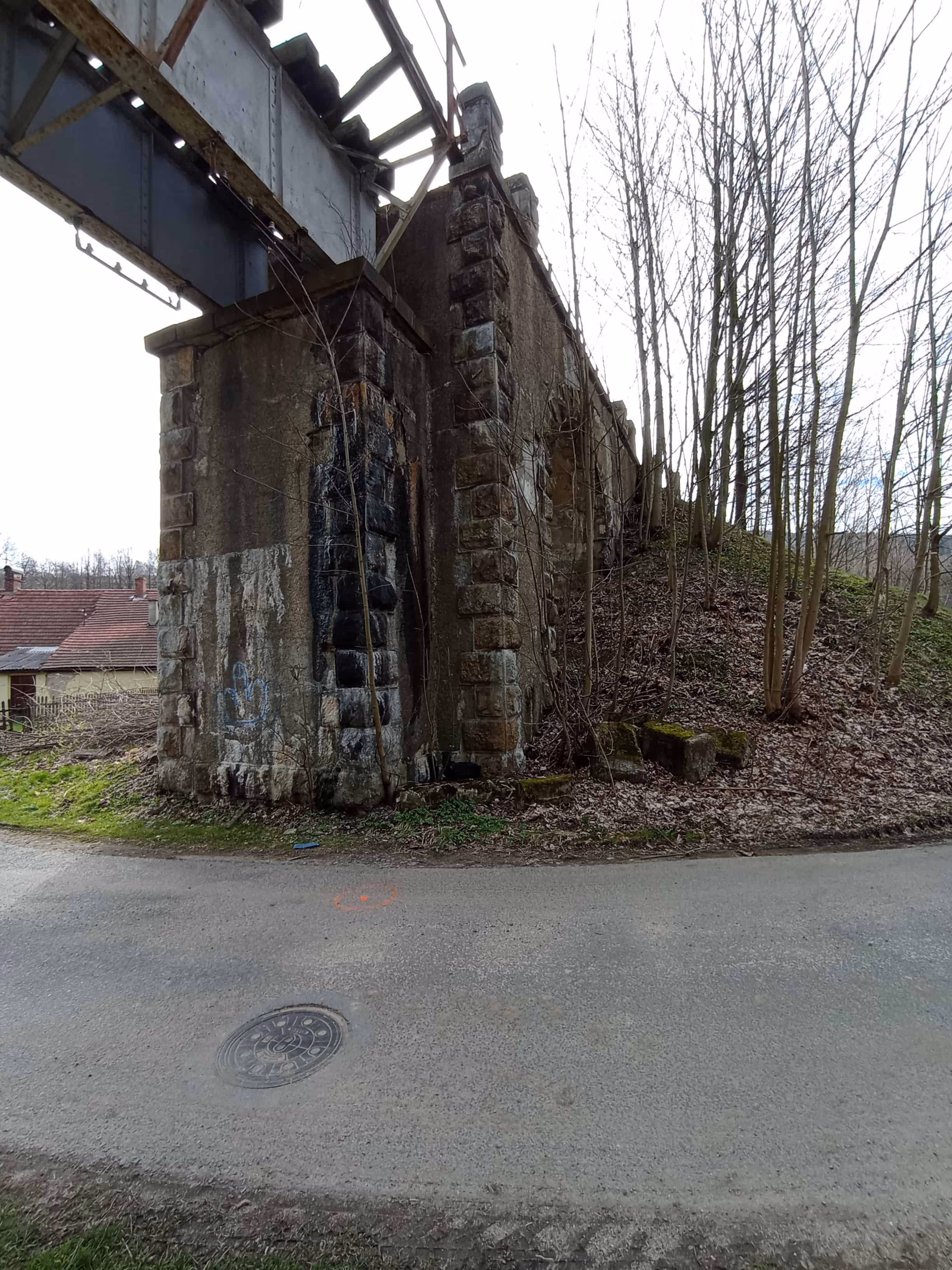 Stary most kolejowy z cegły i betonu nad drogą, otoczony drzewami bez liści i domami z czerwonymi dachami.