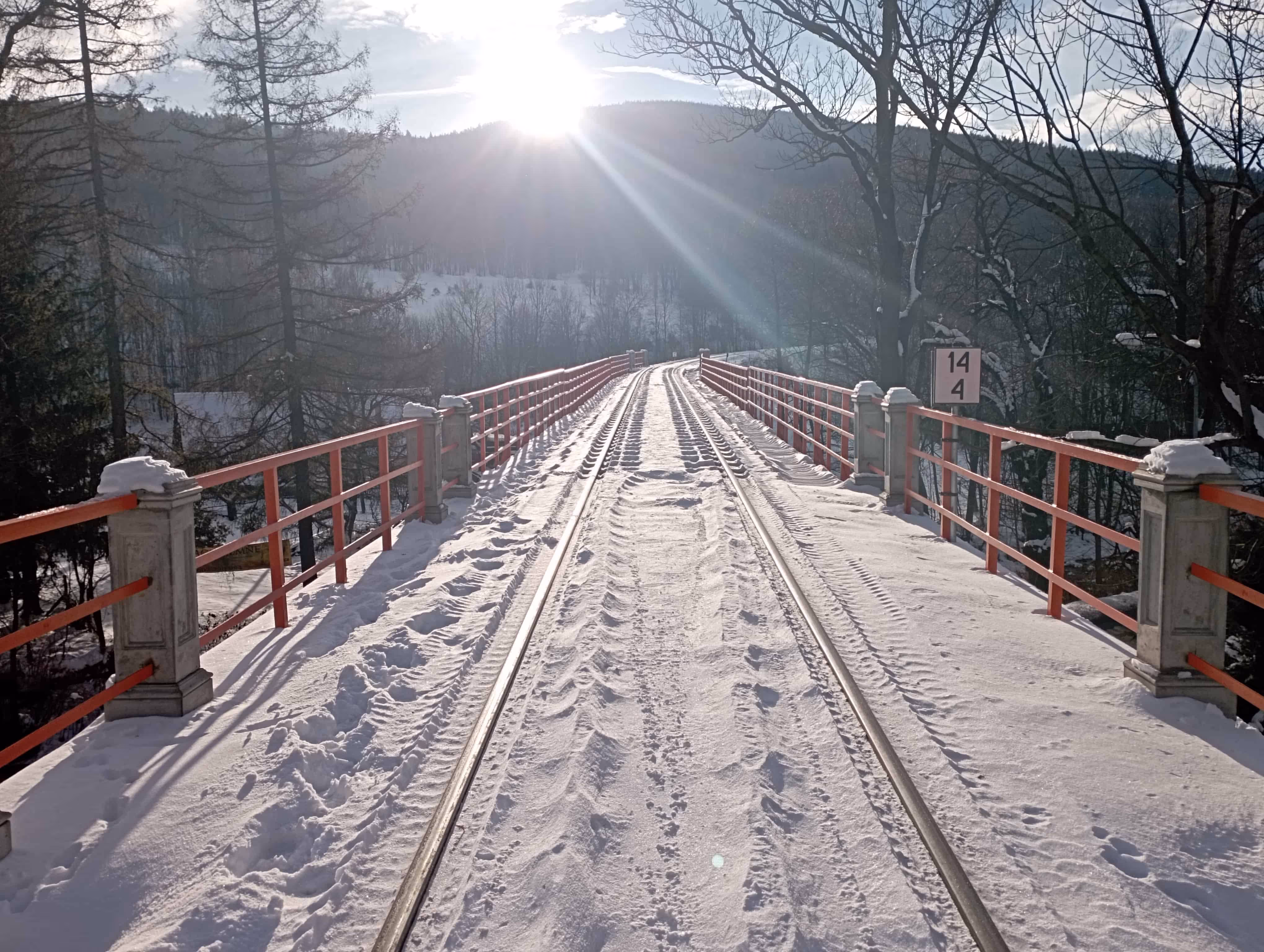 Most kolejowy pokryty śniegiem z czerwonymi poręczami, w tle góry i słońce nisko nad horyzontem.