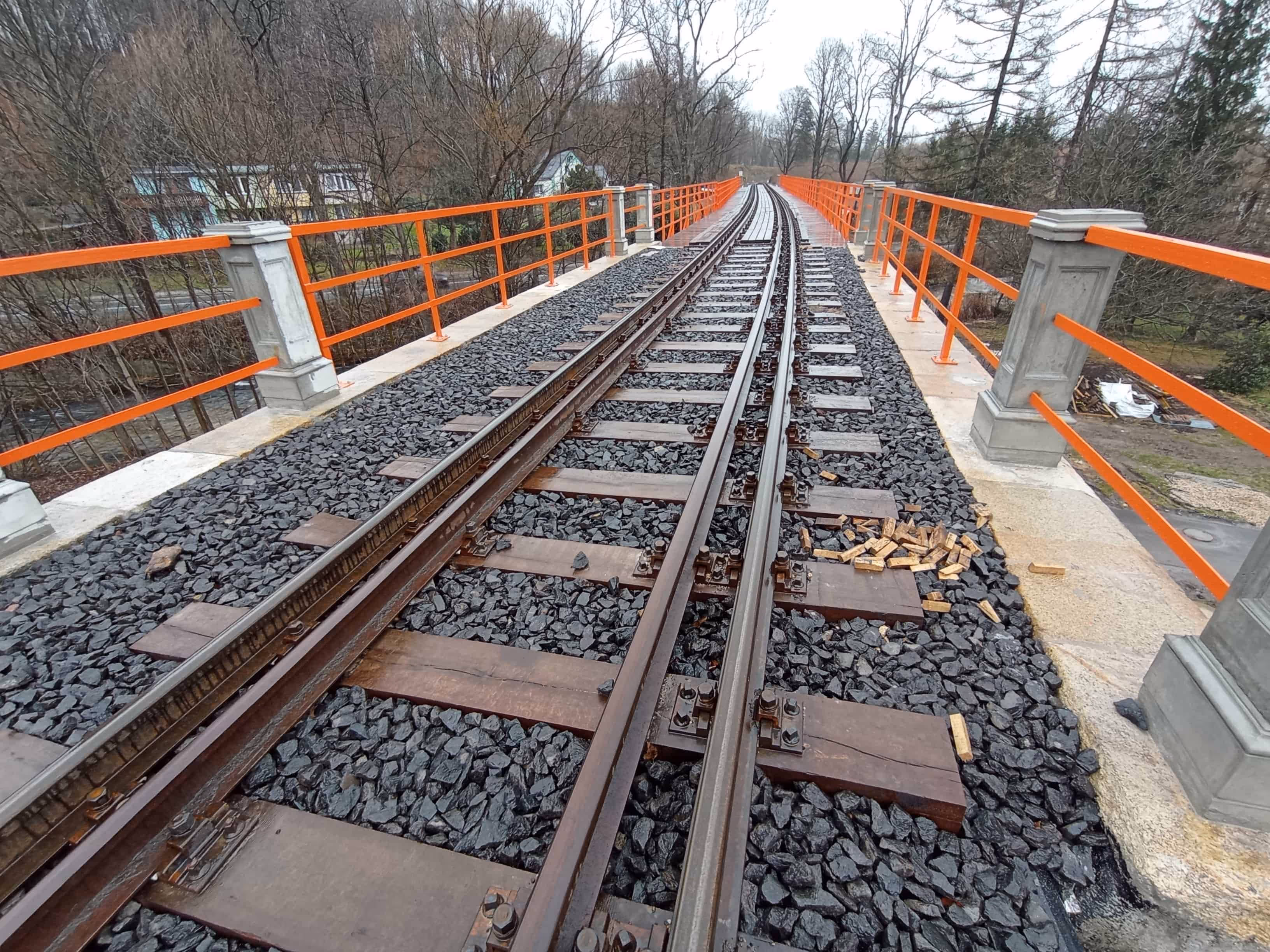 Widok torów kolejowych na moście z pomarańczowymi barierkami, otoczonych drzewami bez liści.