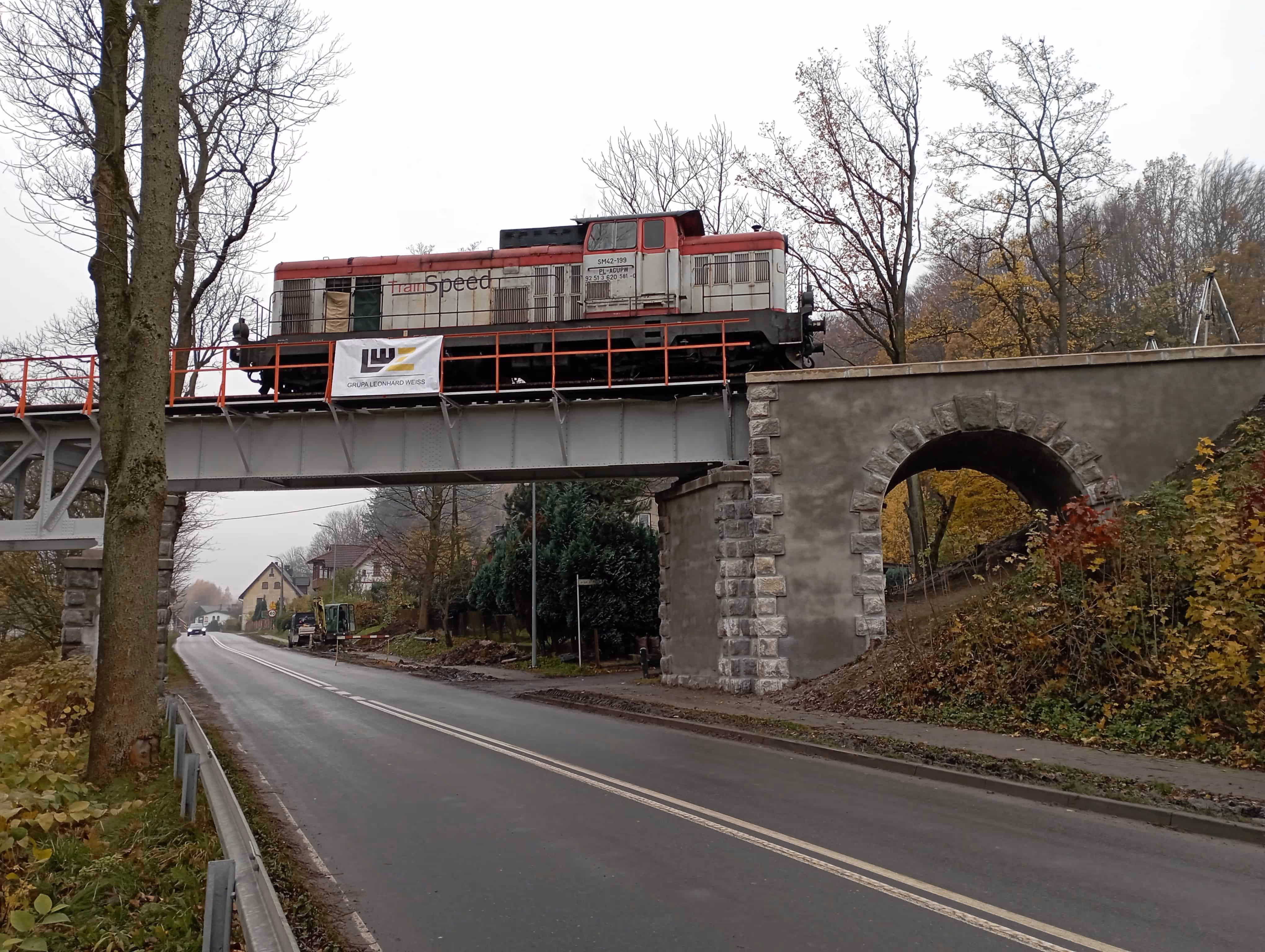 Czerwono-szara lokomotywa stojąca na moście kolejowym nad pustą drogą w jesiennym krajobrazie.