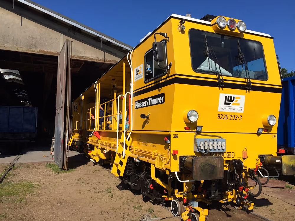 Żółty pojazd kolejowy Plasser & Theurer stojący na torach kolejowych obok budynku, na tle niebieskiego nieba.