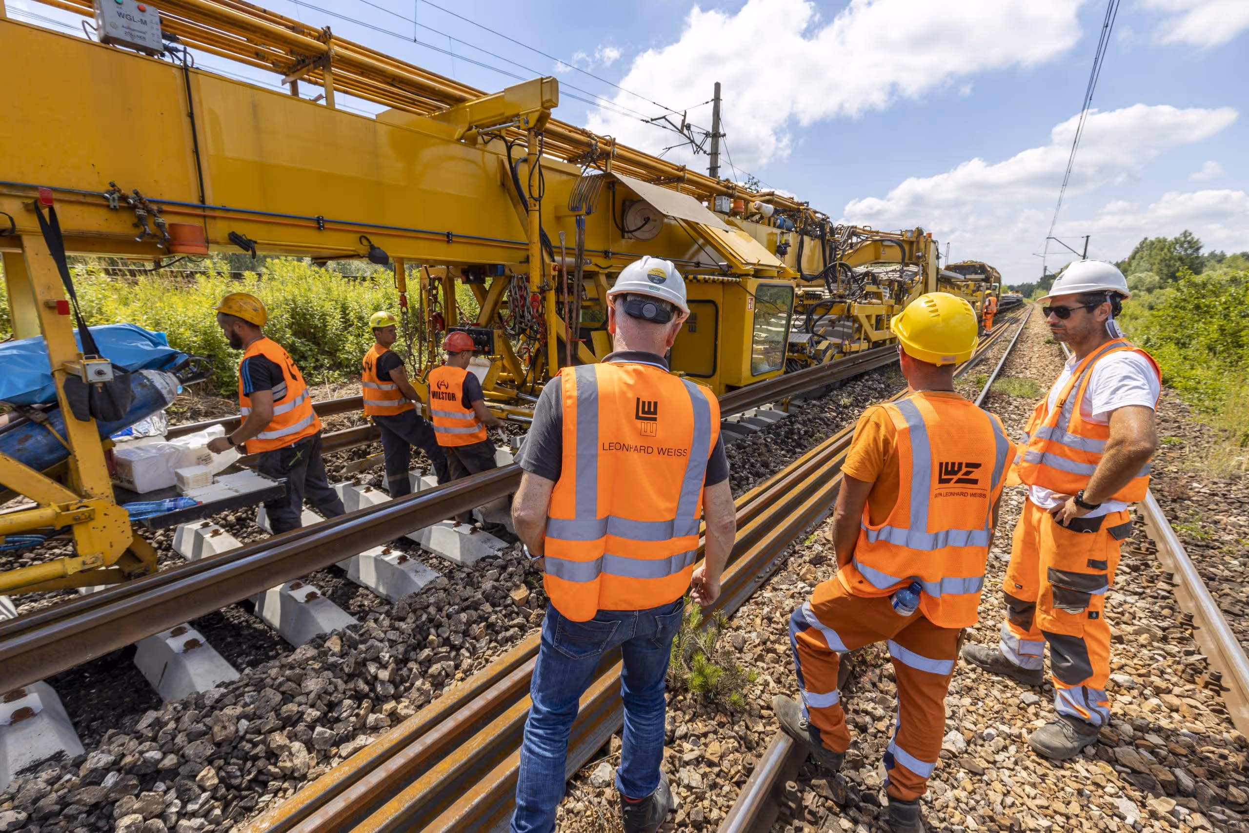 Pracownicy w kamizelkach odblaskowych i kaskach budowlanych nadzorujący maszynę do montażu torów kolejowych na torach.