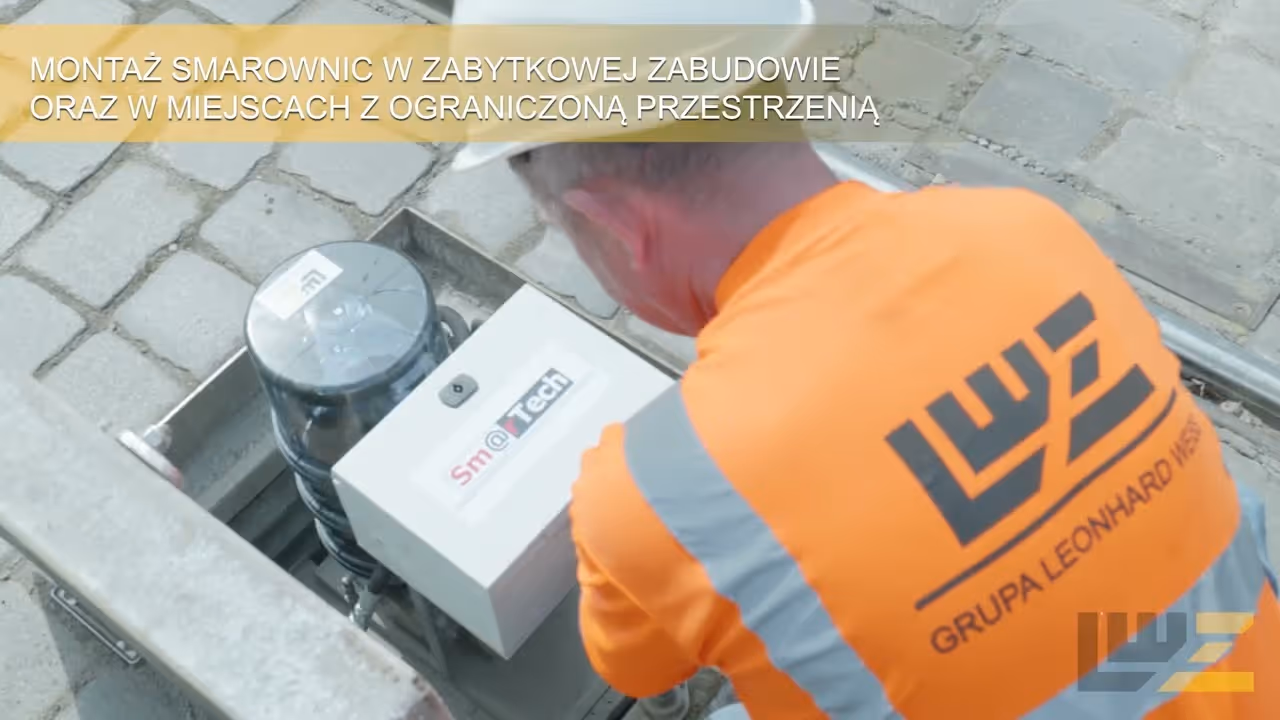 Worker in an orange safety shirt and helmet installing a lubrication device in a small recessed area on a cobblestone street.