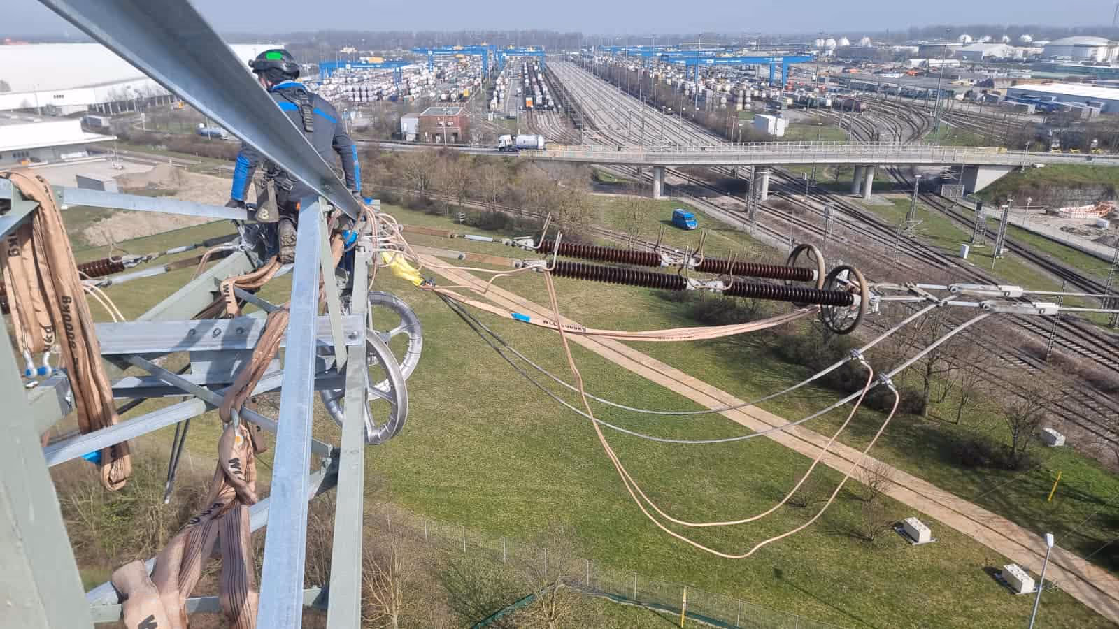 Pracownik konserwujący wysokie linie energetyczne na tle torów kolejowych i zielonego pola.