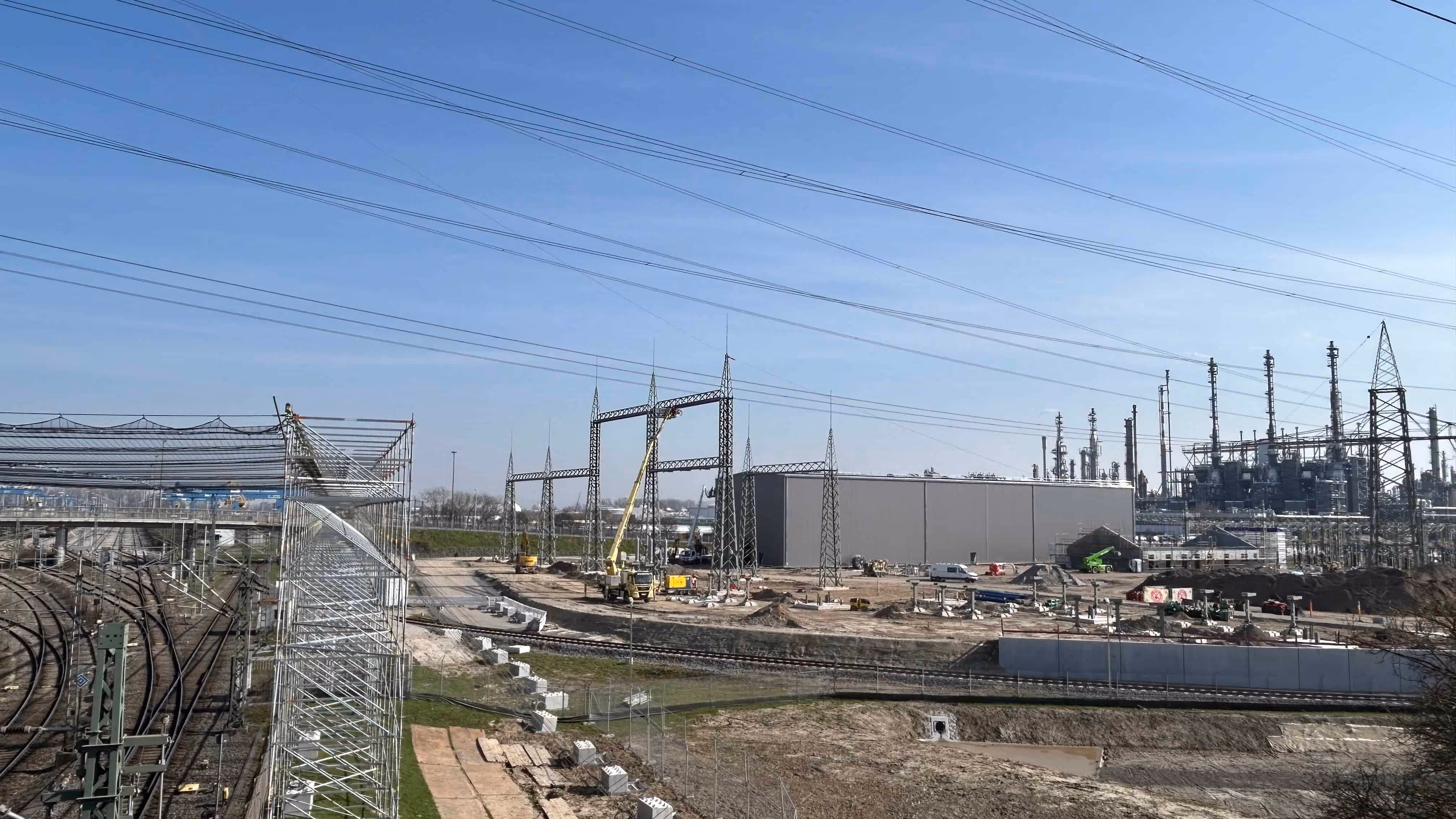 Plac budowy z dużą halą przemysłową, żurawiami i instalacjami energetycznymi na tle nieba.