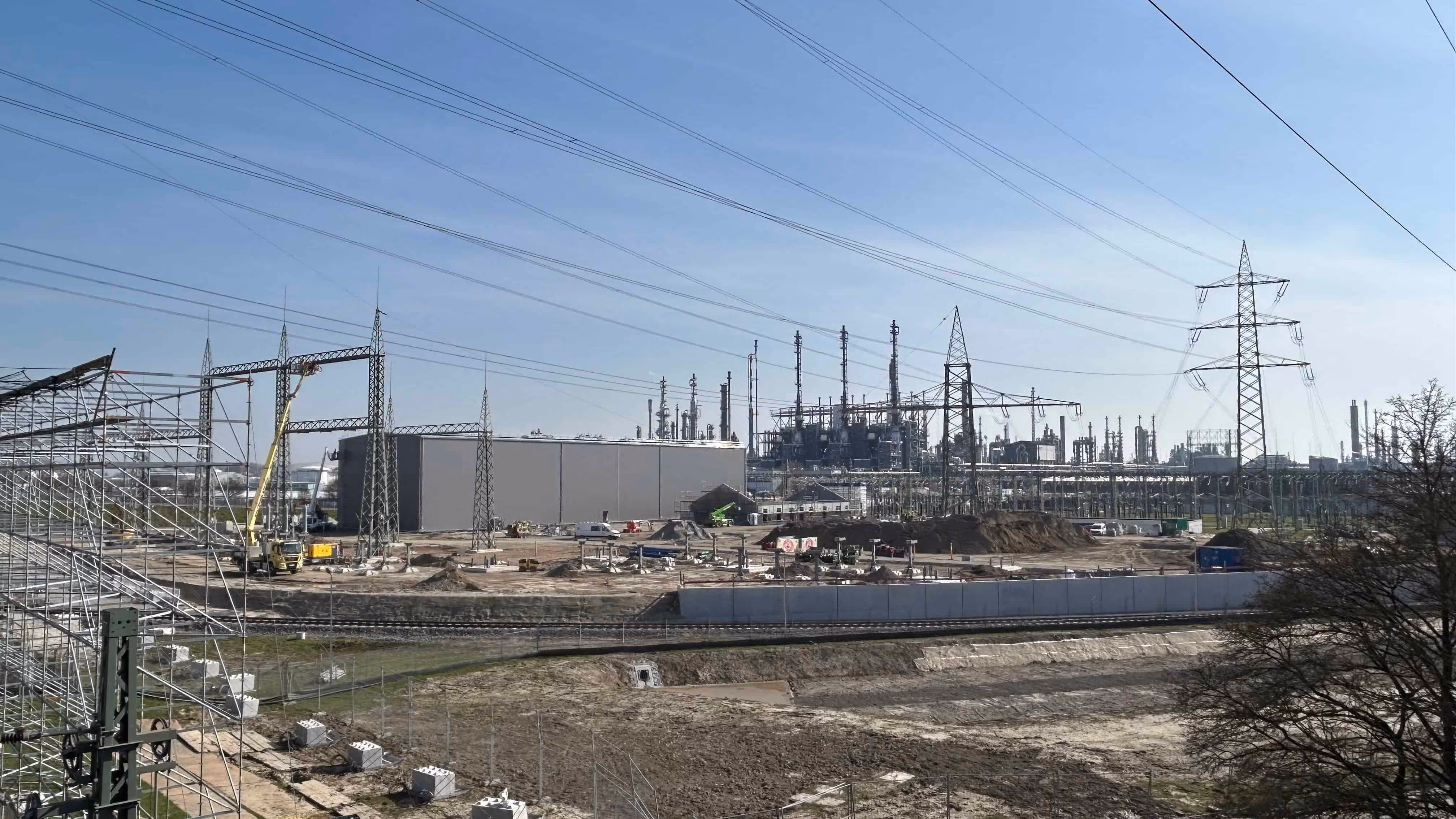 Widok na teren przemysłowy z dużą fabryką, konstrukcjami metalowymi, masztami i słupami wysokiego napięcia na tle niebieskiego nieba.