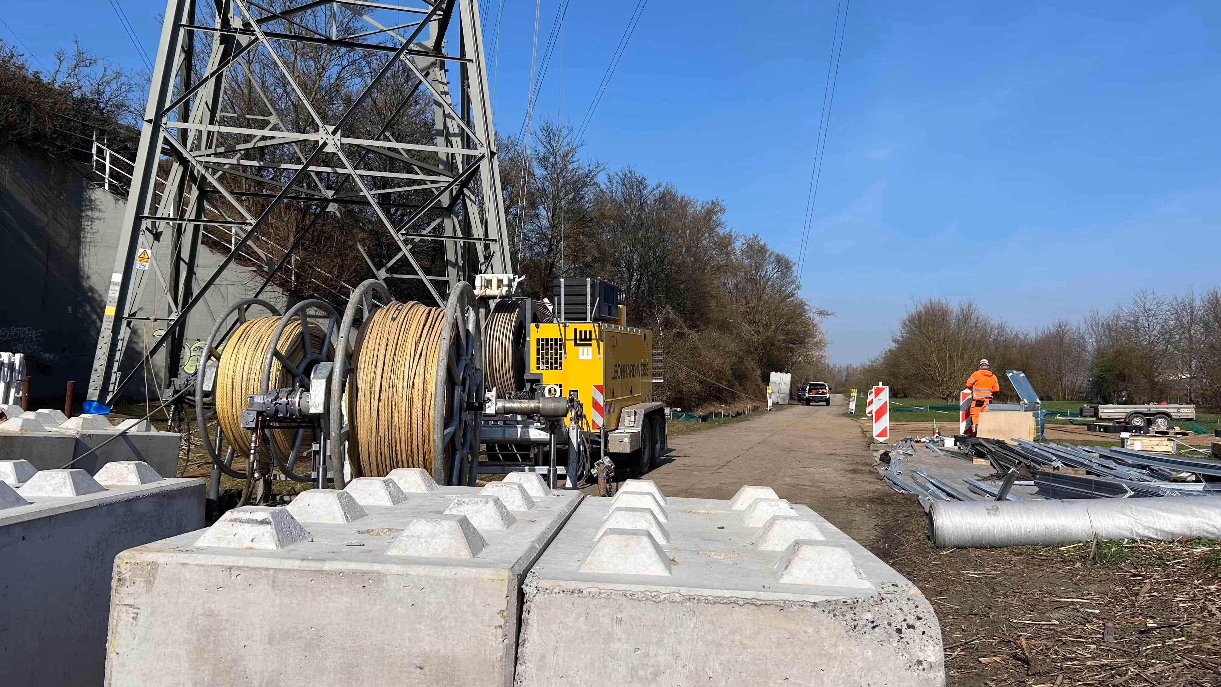 Prace budowlane pod wysokim masztem energetycznym z dużymi bębnami z linami stalowymi i pracownikiem w pomarańczowym ubraniu ochronnym.