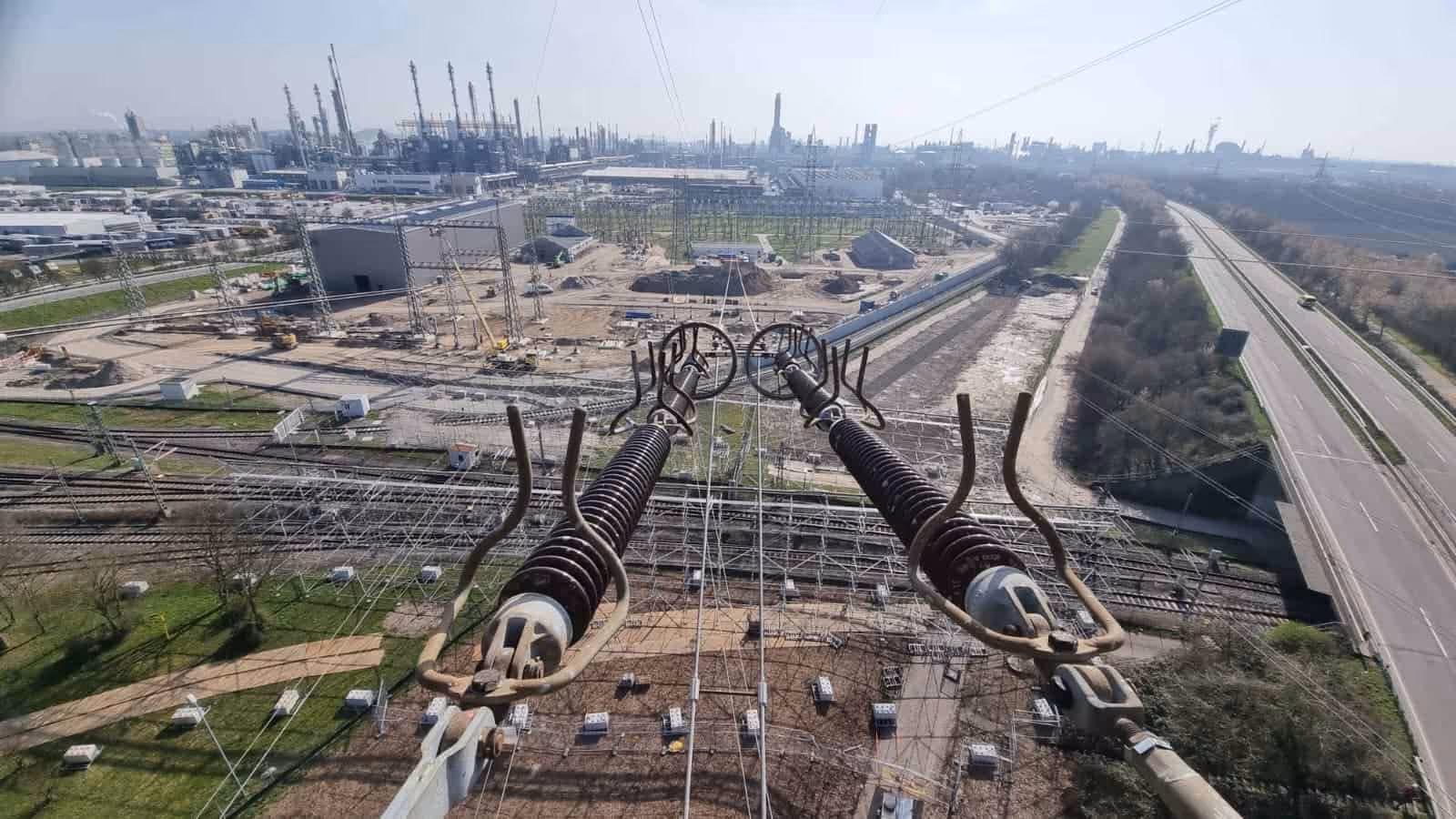 Pomost wysokiego napięcia nad rozległym terenem przemysłowym z instalacjami i liniami kolejowymi.