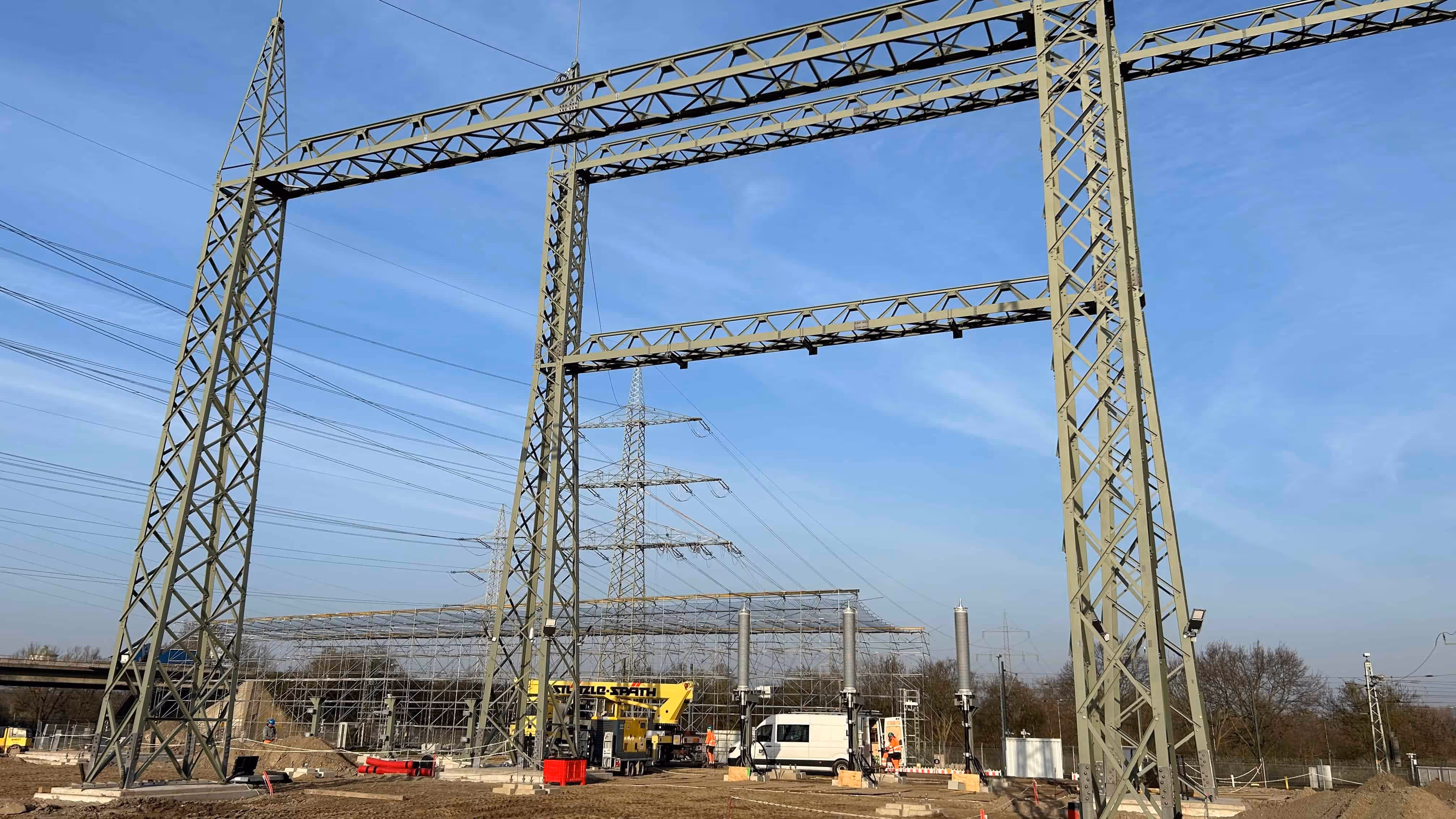 Obiekt przemysłowy z wysokimi metalowymi konstrukcjami i przewodami elektrycznymi na tle niebieskiego nieba.