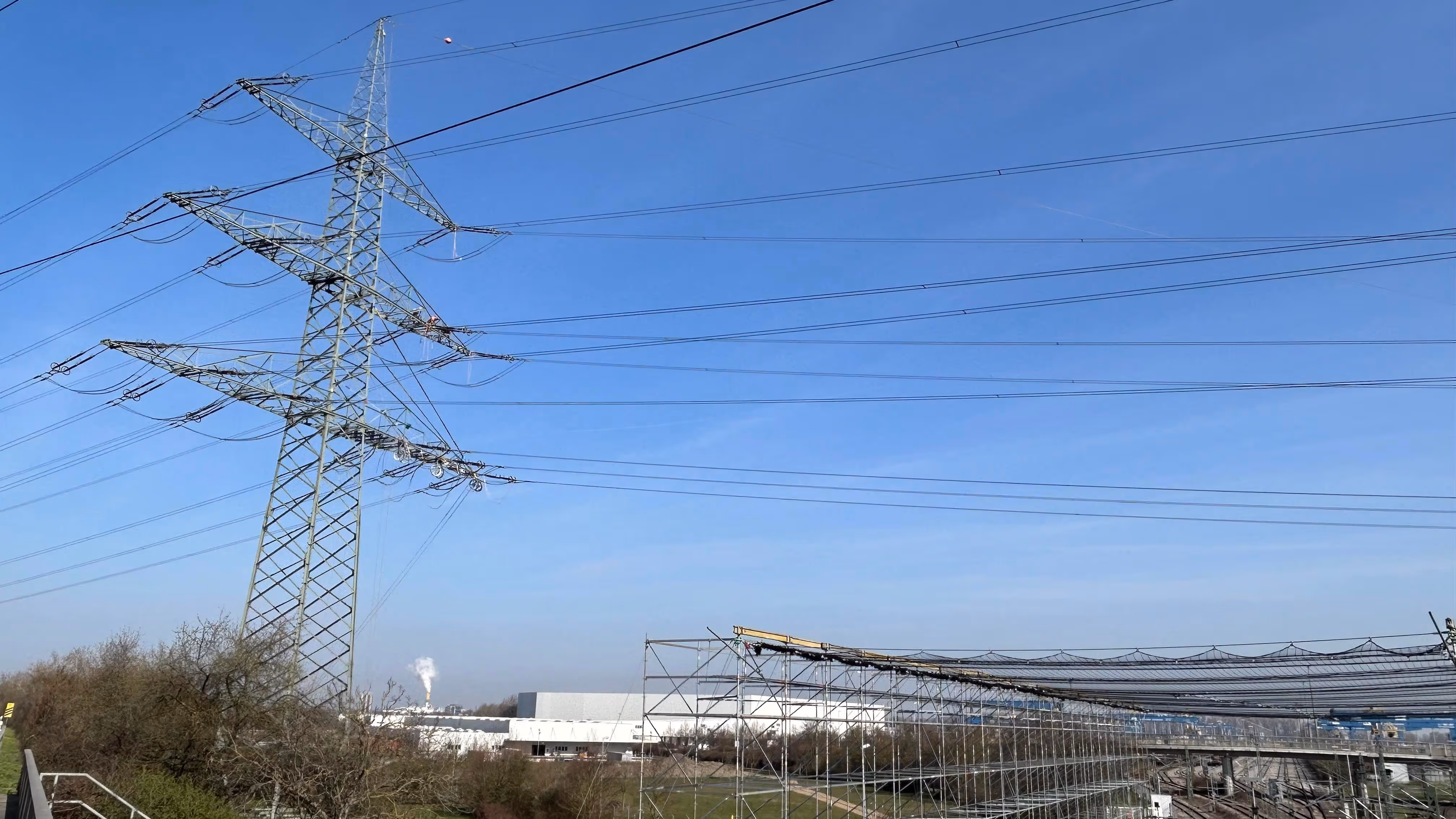 Metal electricity pylon with multiple power lines against a clear blue sky over industrial buildings and bushes.