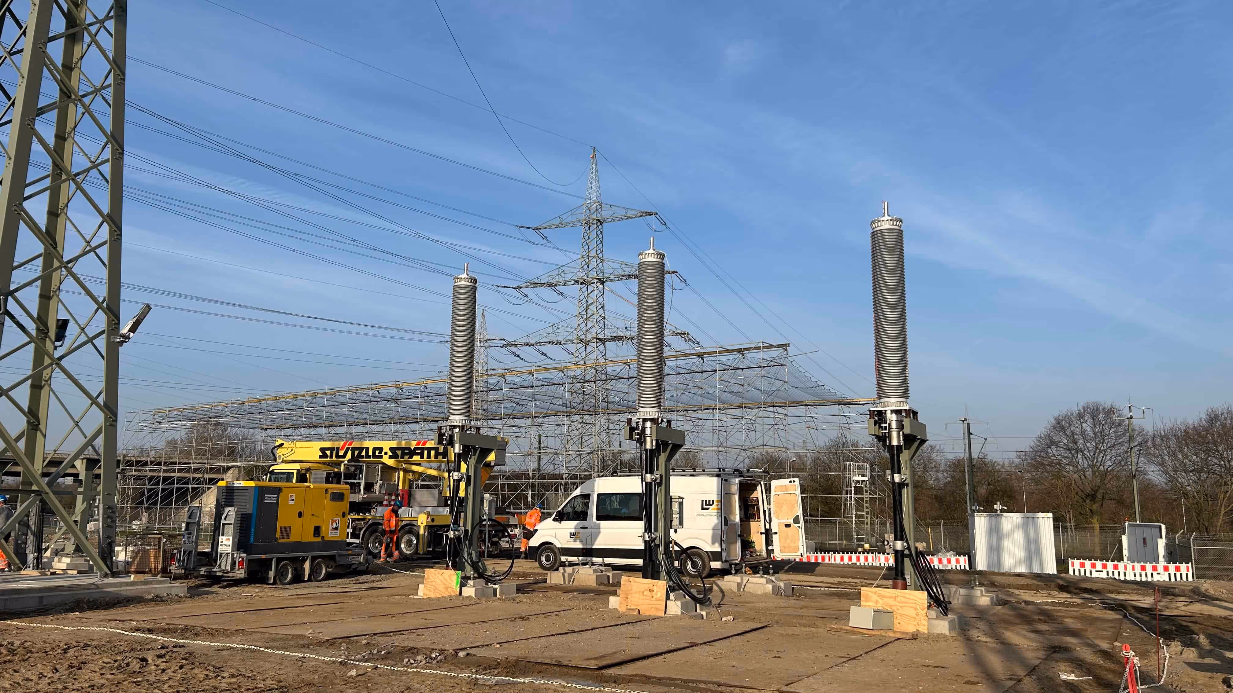 Plac budowy stacji elektrycznej z trzema dużymi izolatorami i pracownikami przy sprzęcie budowlanym.