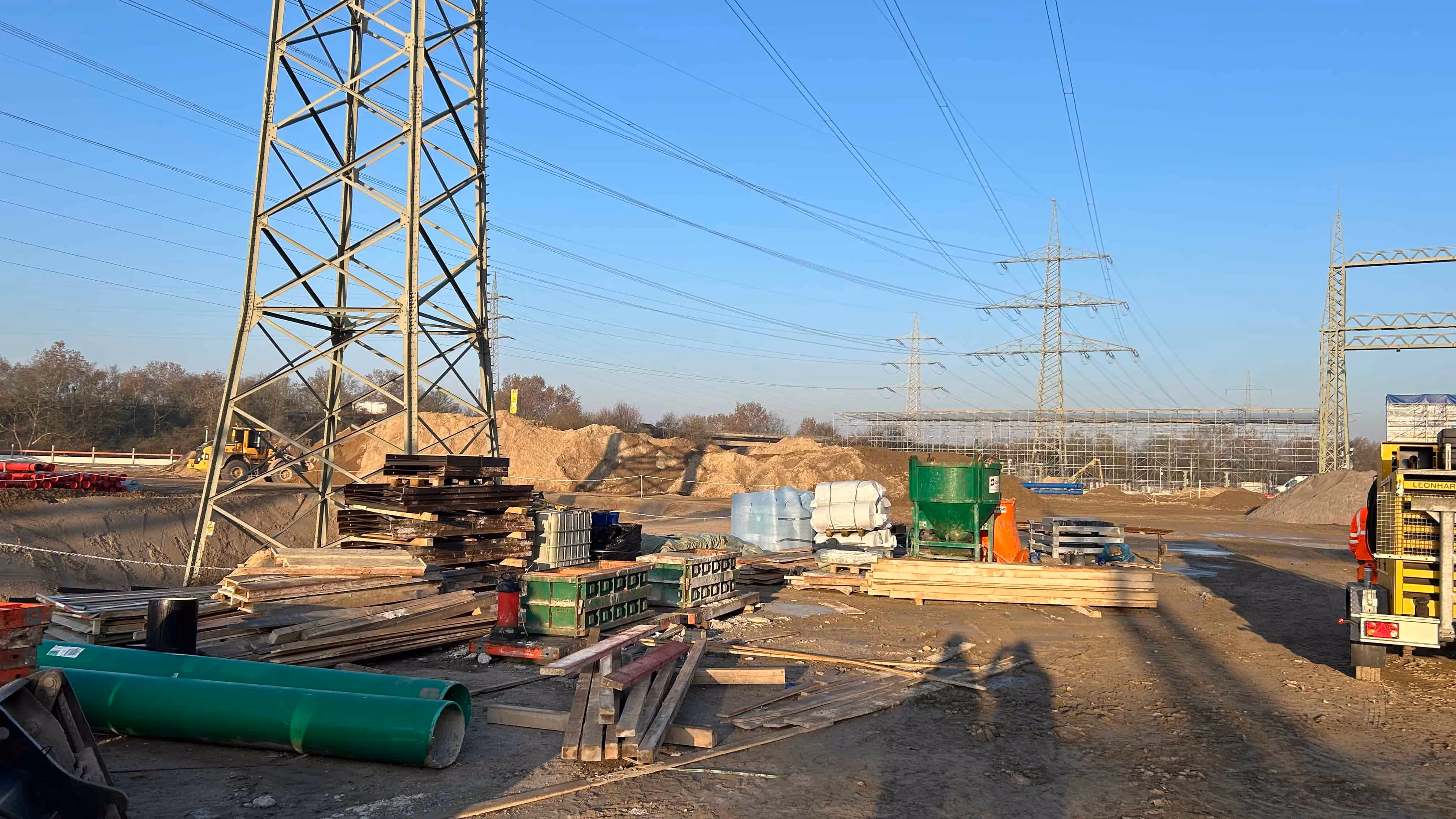 Plac budowy z narzędziami, drewnem i materiałami na ziemi pod liniami wysokiego napięcia na tle niebieskiego nieba.