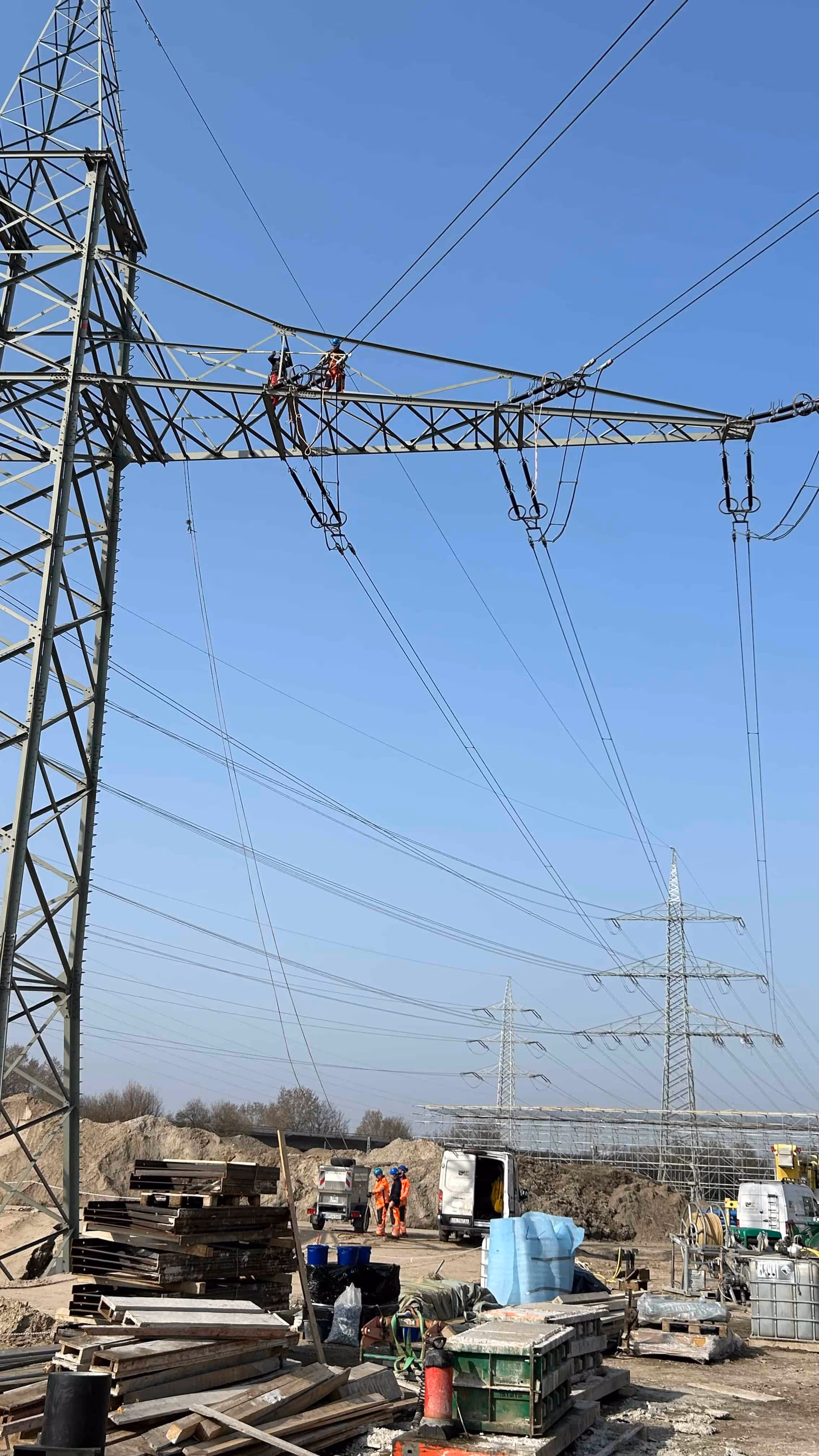 Pracownik na wieży energetycznej wykonujący prace na wysokości, z grupą robotników i sprzętem budowlanym na ziemi pod nim.
