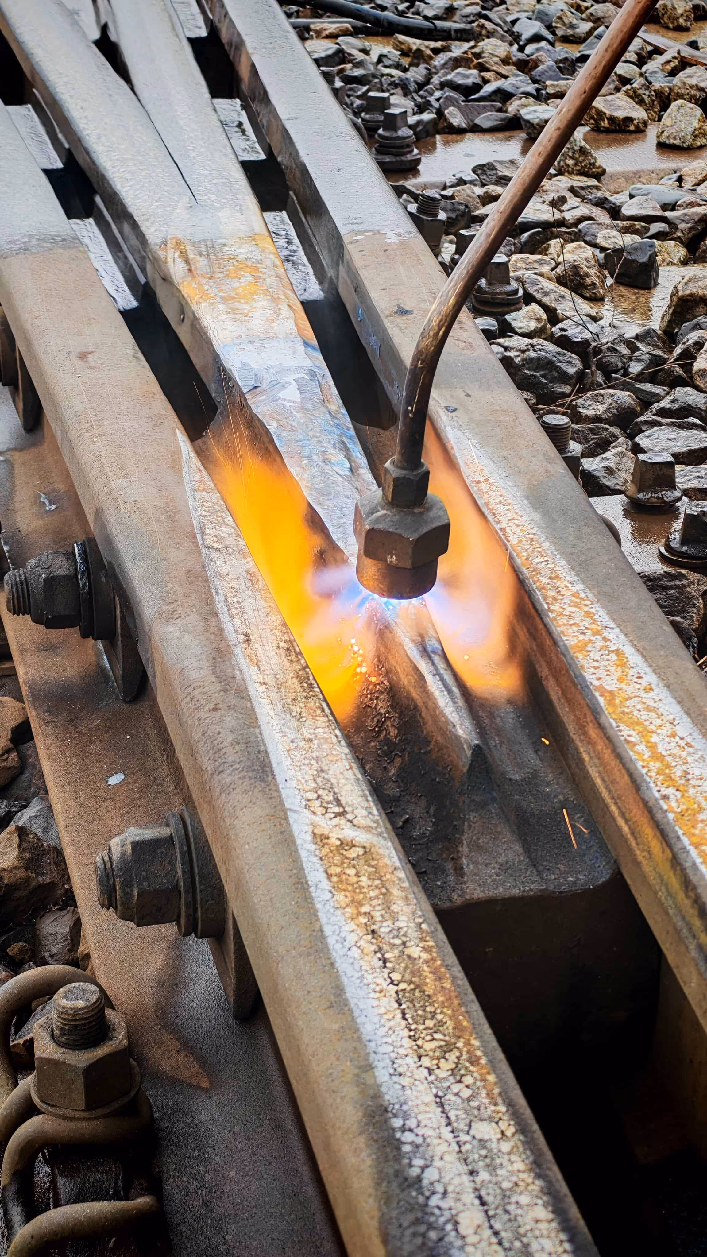 Palnik gazowy podgrzewający metalowy szynę kolejową na tle żwiru i podkładów.