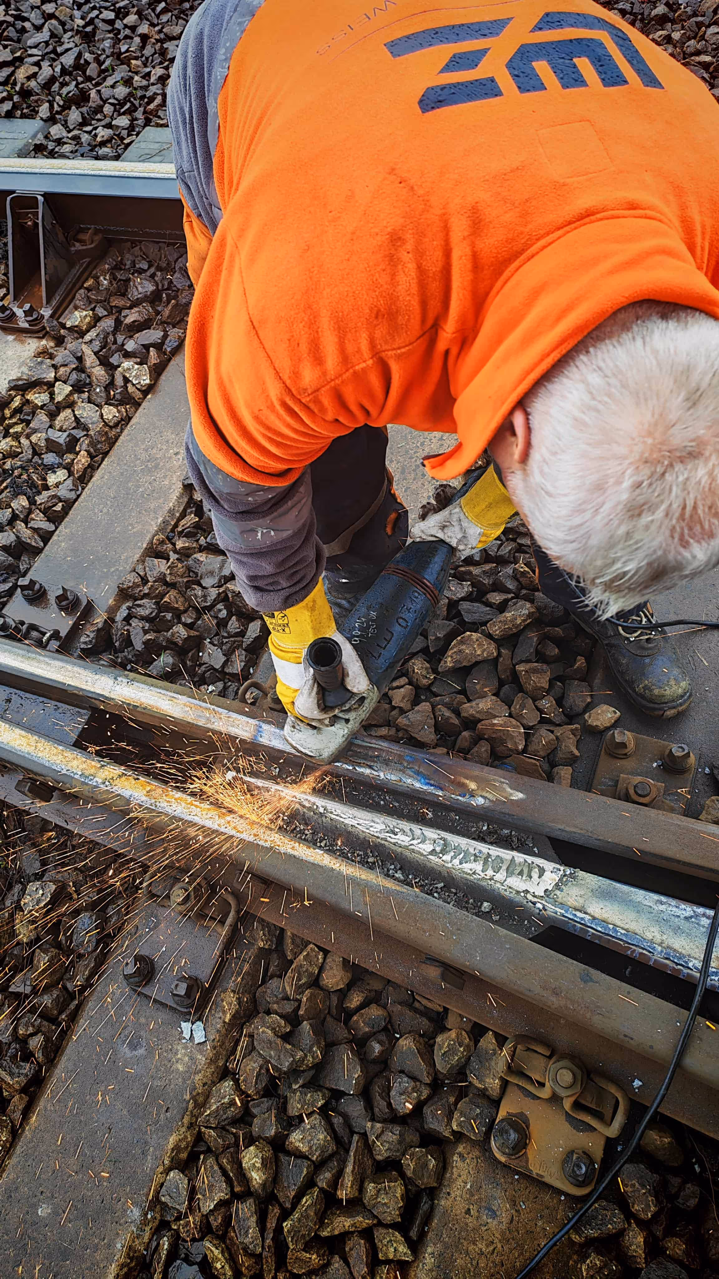 Robotnik w pomarańczowej kurtce szlifuje metalową szynę kolejową, iskrząc podczas pracy.