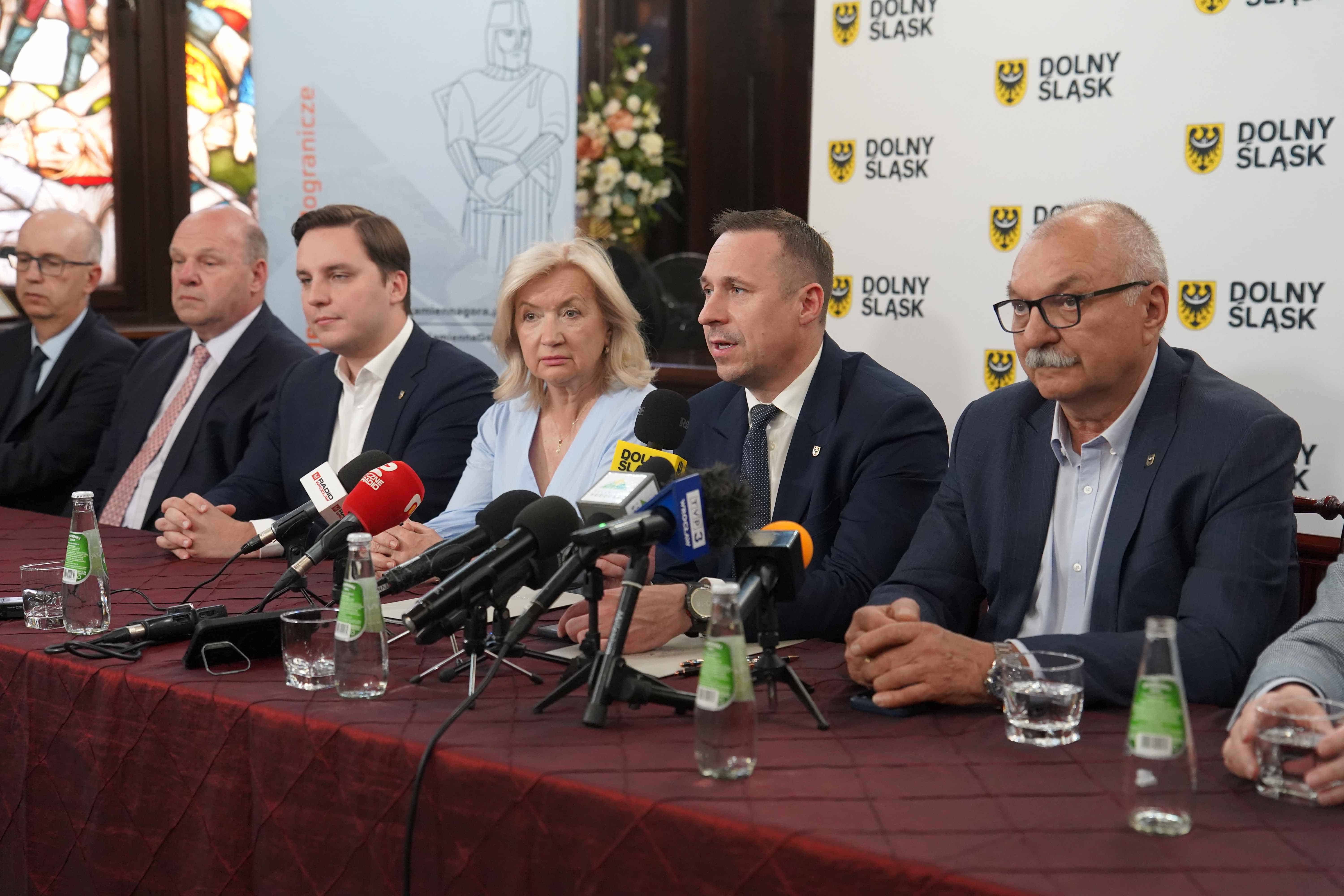 Grupa sześciu osób siedzi przy stole podczas konferencji z mikrofonami i butelkami wody.