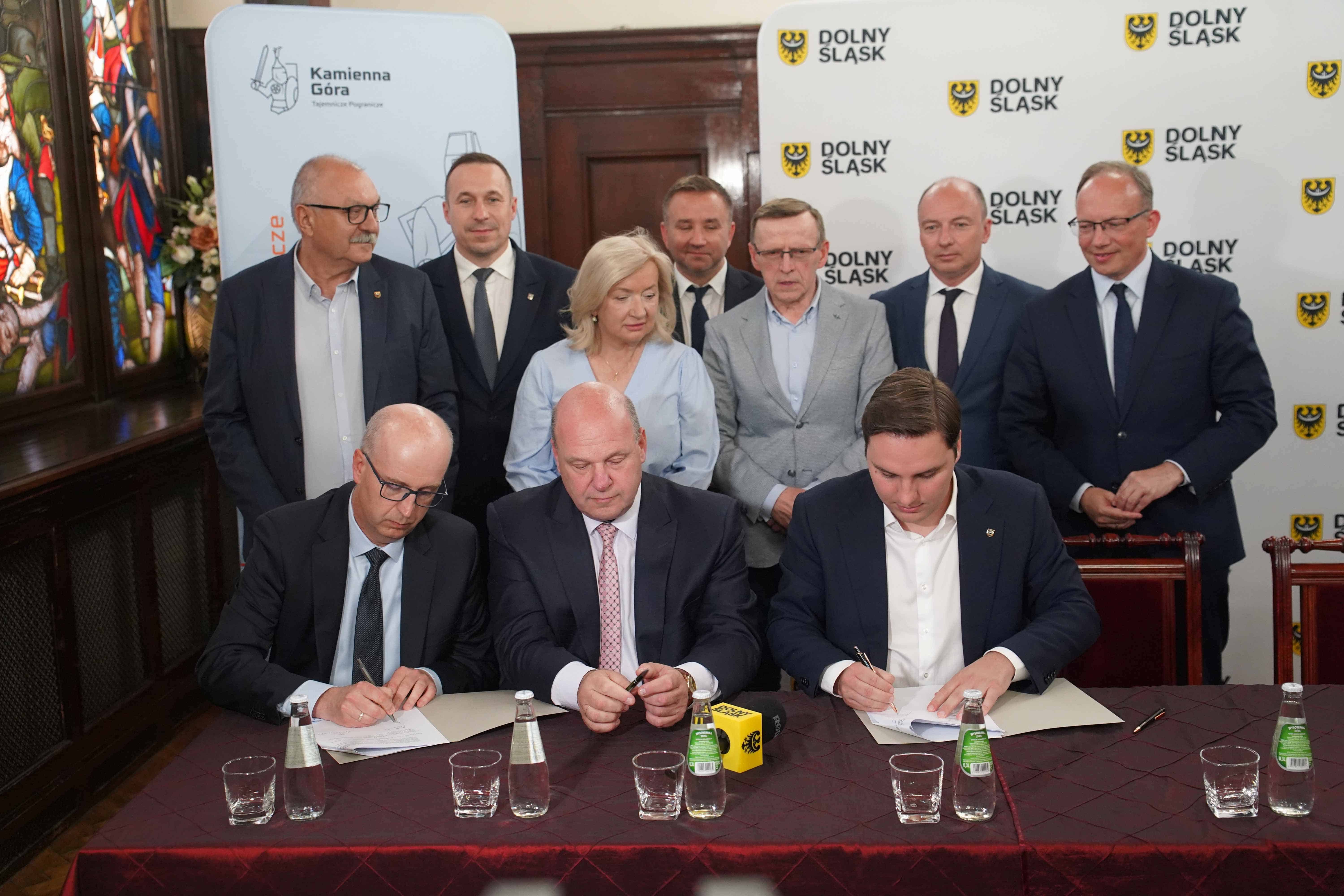 Trzech mężczyzn podpisuje dokumenty przy stole, z grupą osób stojących za nimi w oficjalnym pomieszczeniu.