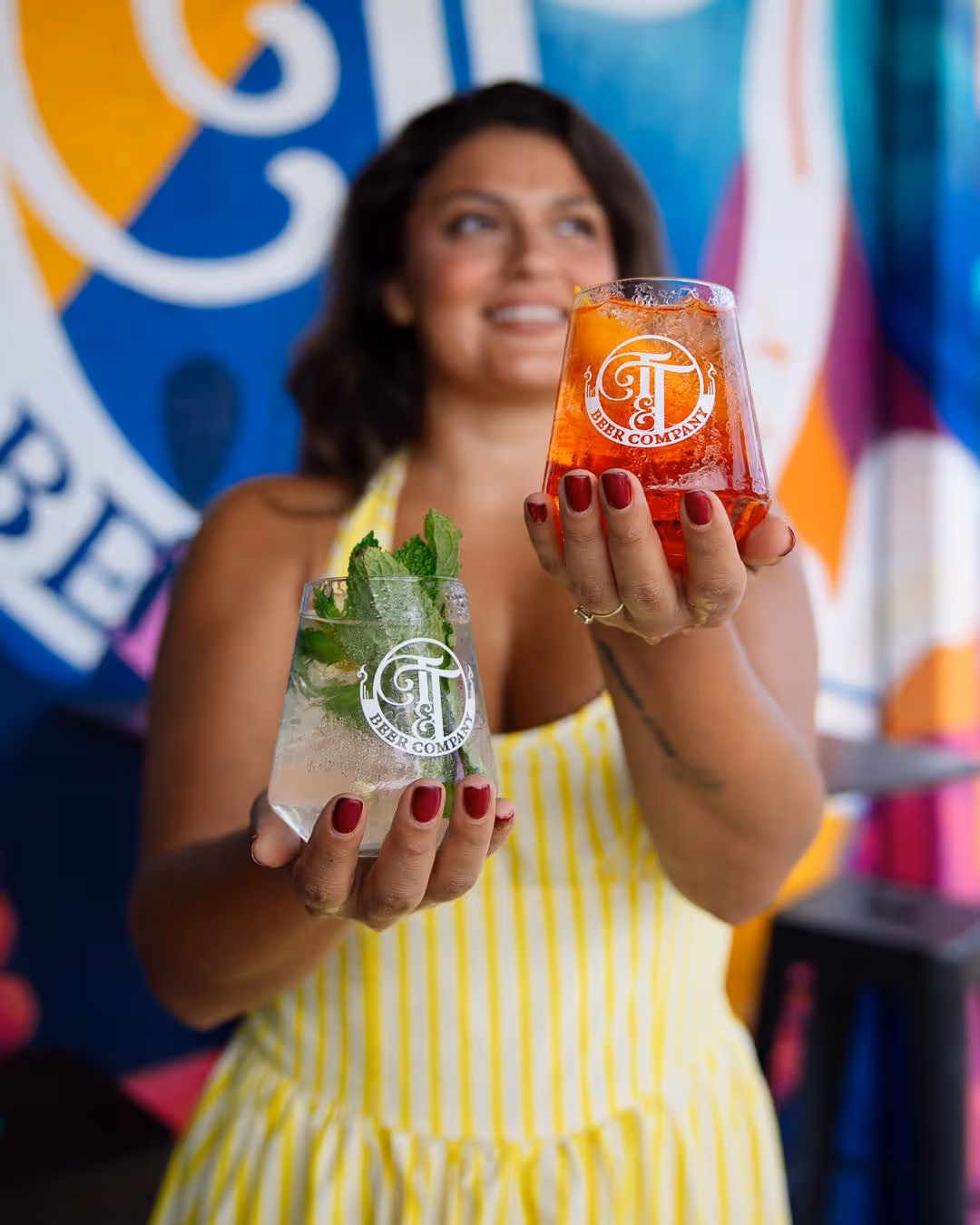 Woman in yellow striped dress holding two branded glasses with cocktails, one garnished with mint and the other with an orange slice.
