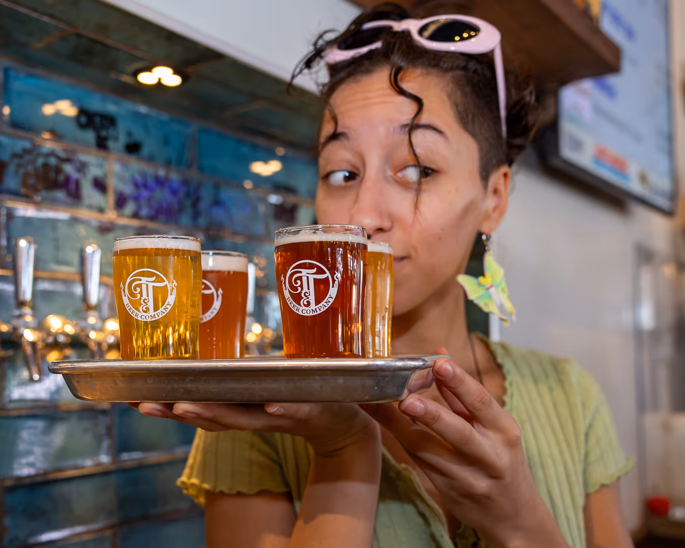 Woman holding a tray with four small glasses of craft beer, each labeled with a beer company logo, inside a brewery.