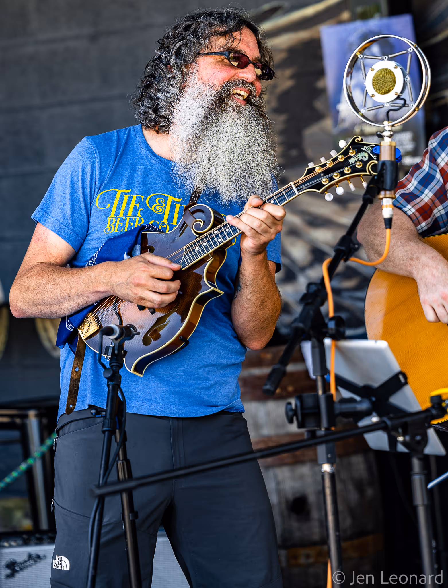 Smiling man with long gray beard playing a mandolin and singing into a vintage microphone.