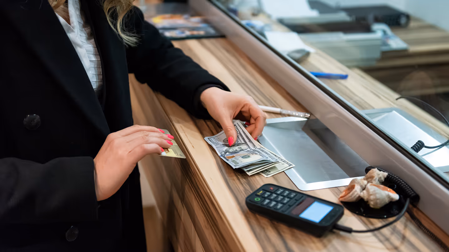 Person with painted nails holding cash and a card at a bank teller window with a card reader on the counter.