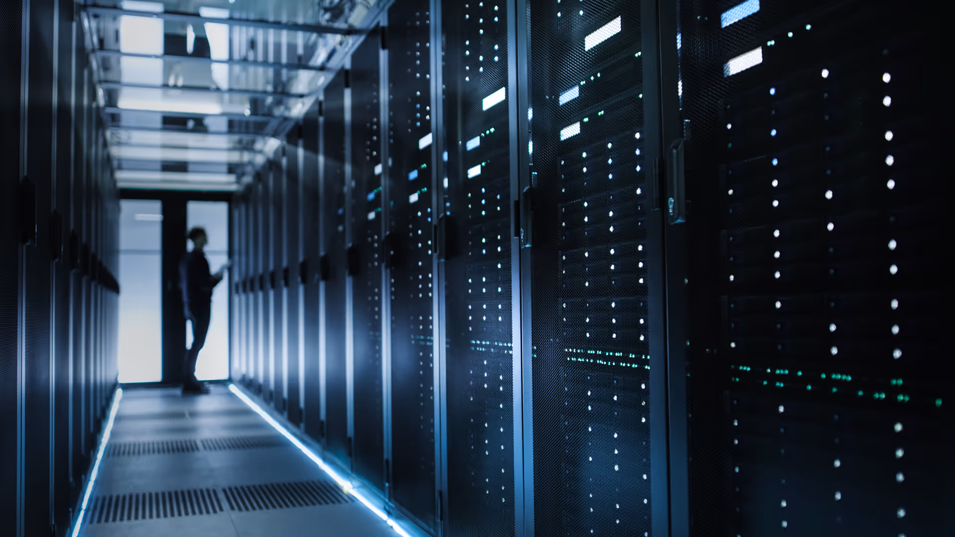 Person standing at the end of a dimly lit data center aisle with server racks illuminated by blue lights.