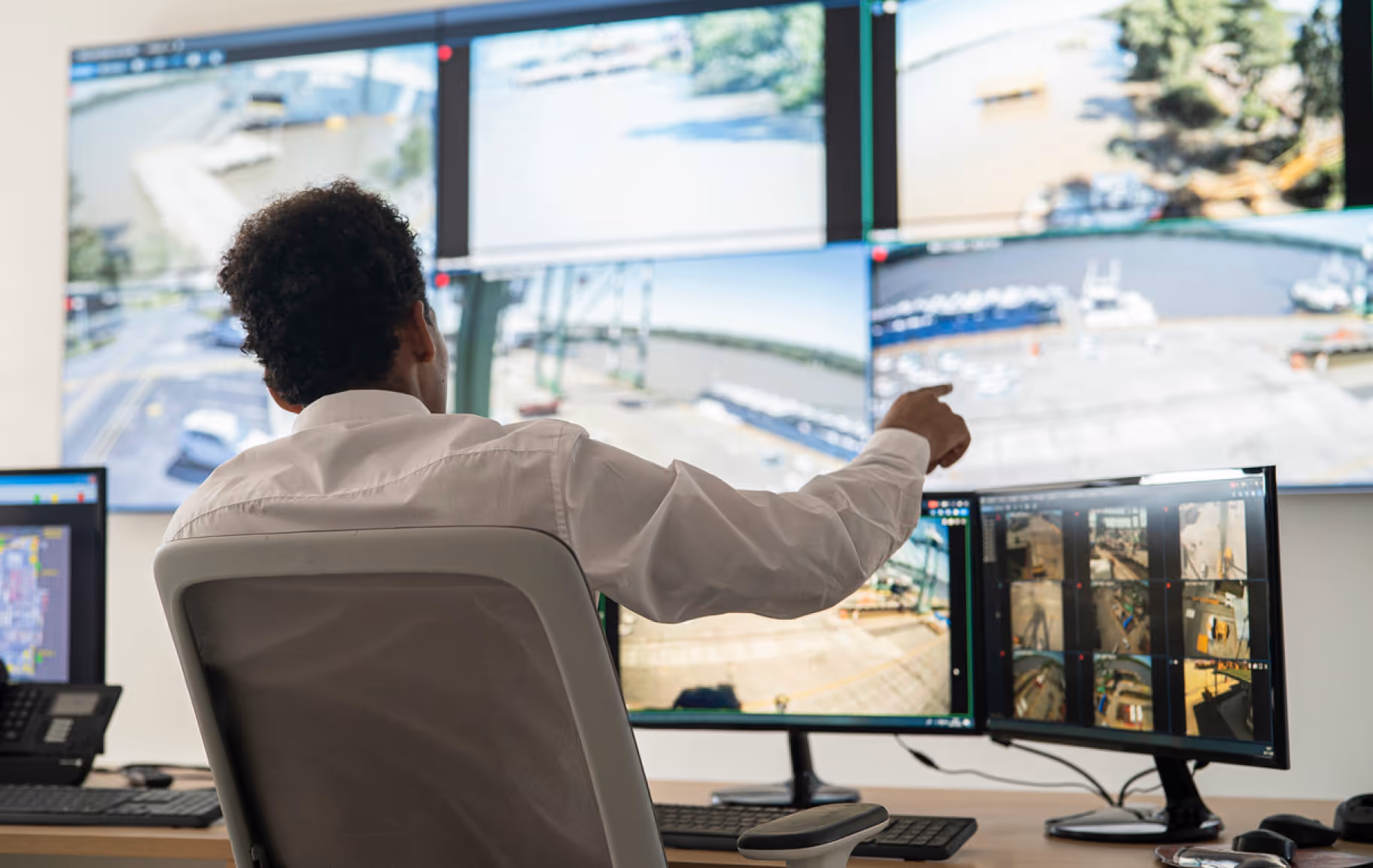 Man in white shirt sitting in front of multiple monitors displaying surveillance footage of outdoor industrial and port areas.