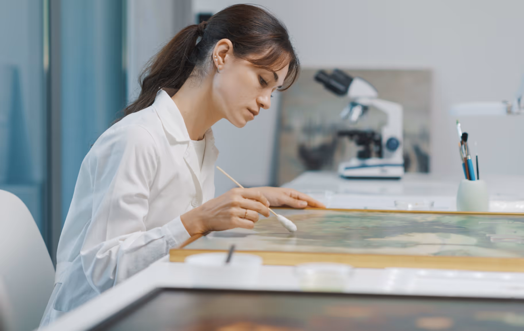 Woman in a lab coat carefully cleaning a framed artwork with a cotton swab in a restoration studio.