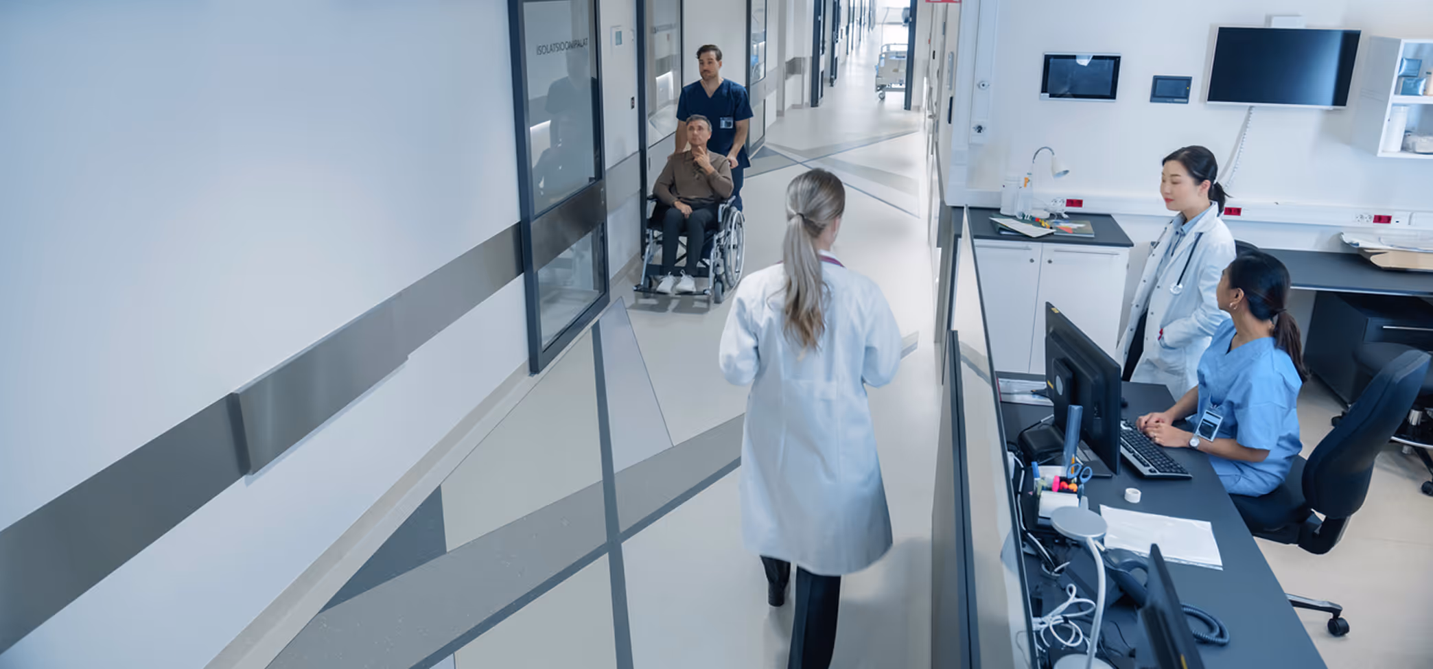 Male nurse pushing an elderly male patient in a wheelchair along a hospital corridor past a nurse station with two female medical staff.