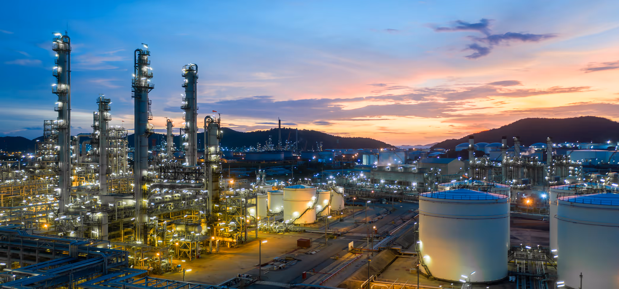 Large industrial oil refinery facility with tall distillation towers and storage tanks illuminated at dusk with mountains in the background.