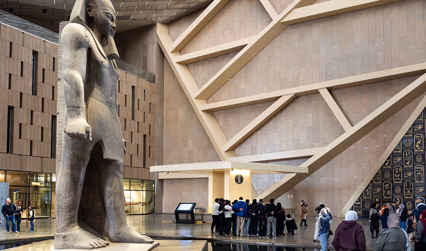 Large ancient Egyptian statue in a modern museum hall with visitors gathered around and architectural ceiling beams.