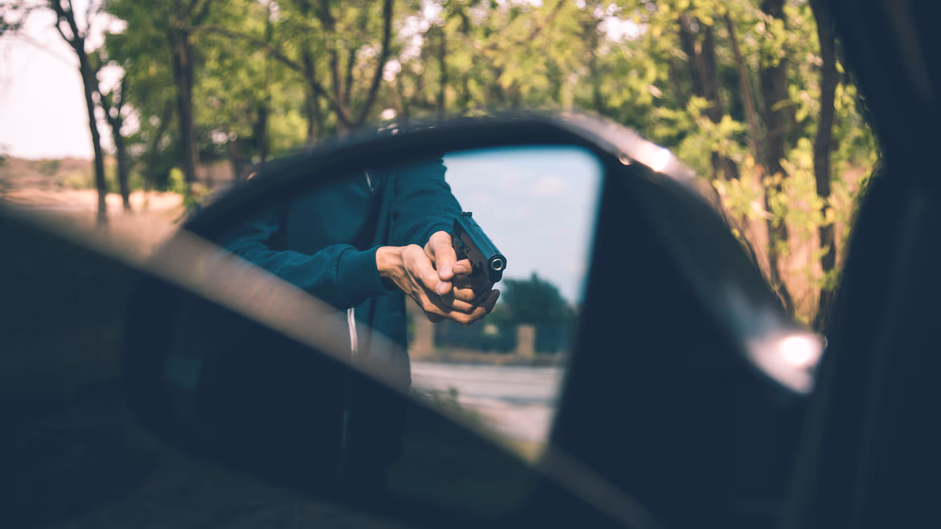 Person aiming a handgun reflected in a car's side mirror with trees in the background.