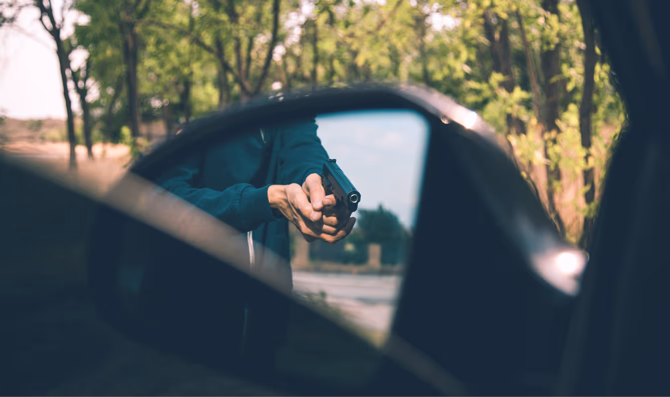 Person aiming a handgun reflected in a car's side mirror with trees in the background.