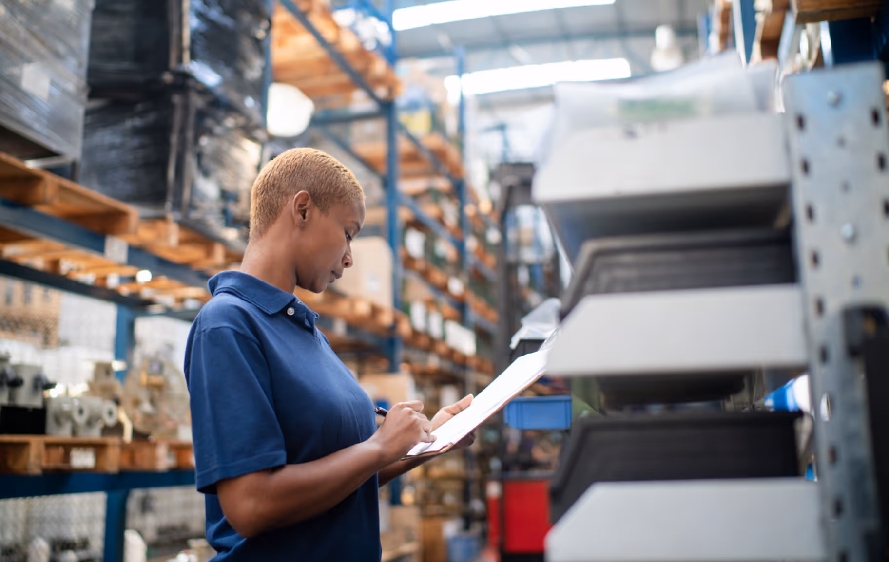Warehouse worker in a blue polo shirt writing on a clipboard amid stacked inventory shelves.