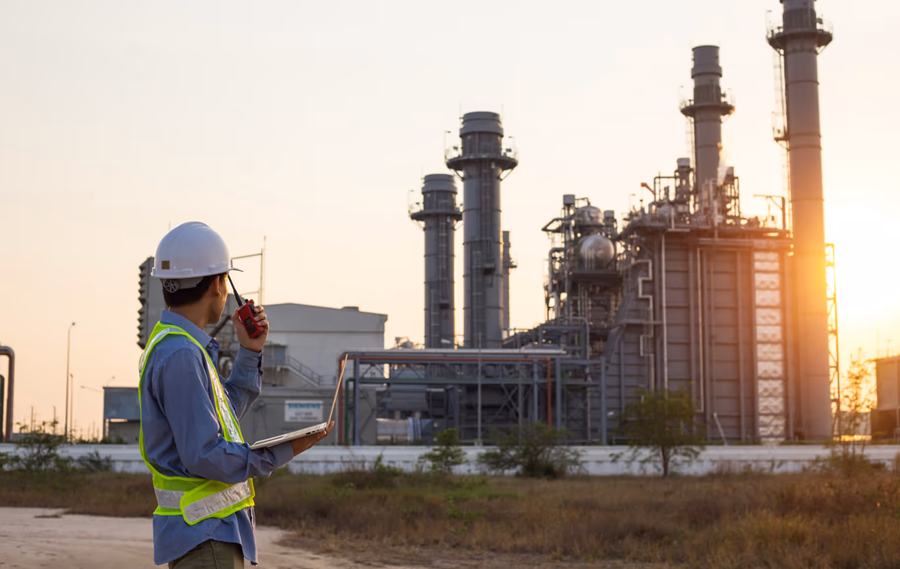 Engineer in a hard hat and safety vest holding a laptop and walkie-talkie at an industrial power plant during sunset.