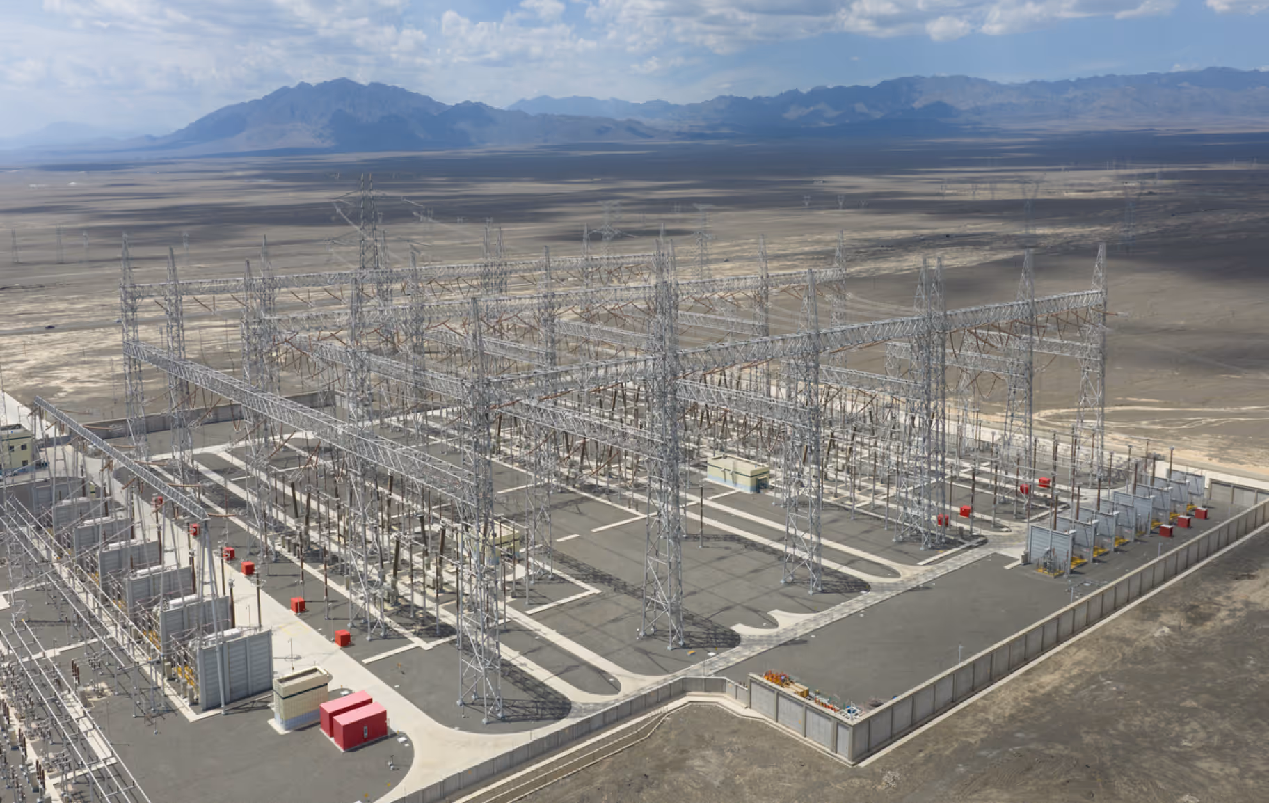 Aerial view of a large electrical substation with metal towers and power lines in a desert landscape with mountains in the background.