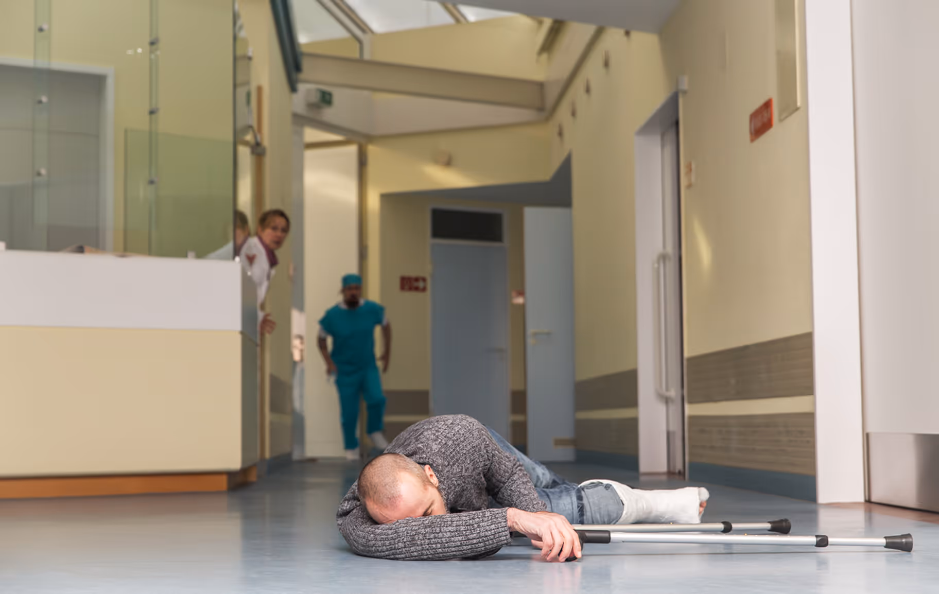 Man with a leg cast lying on the hospital floor holding crutches, medical staff rushing toward him.