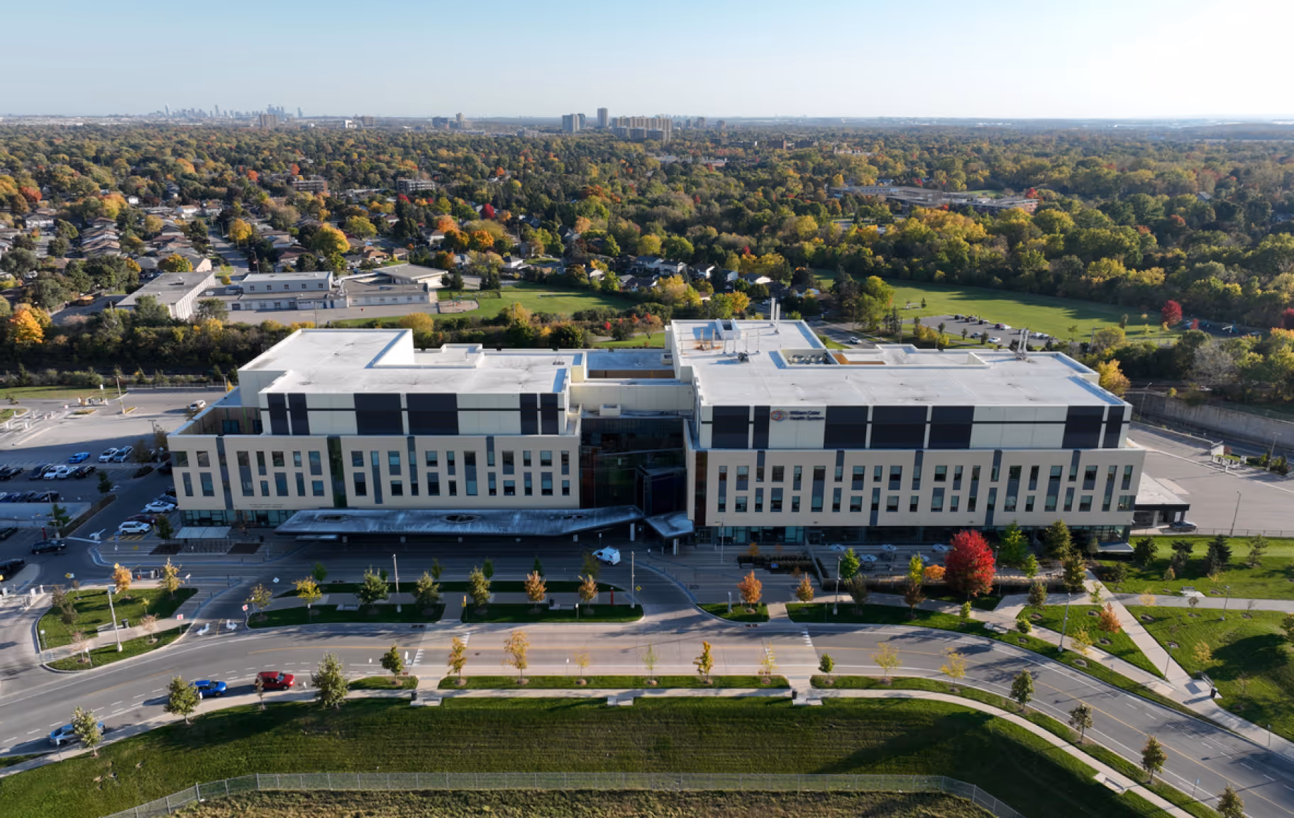 Aerial view of a large modern hospital building surrounded by roads, trees, and green spaces in a suburban area.