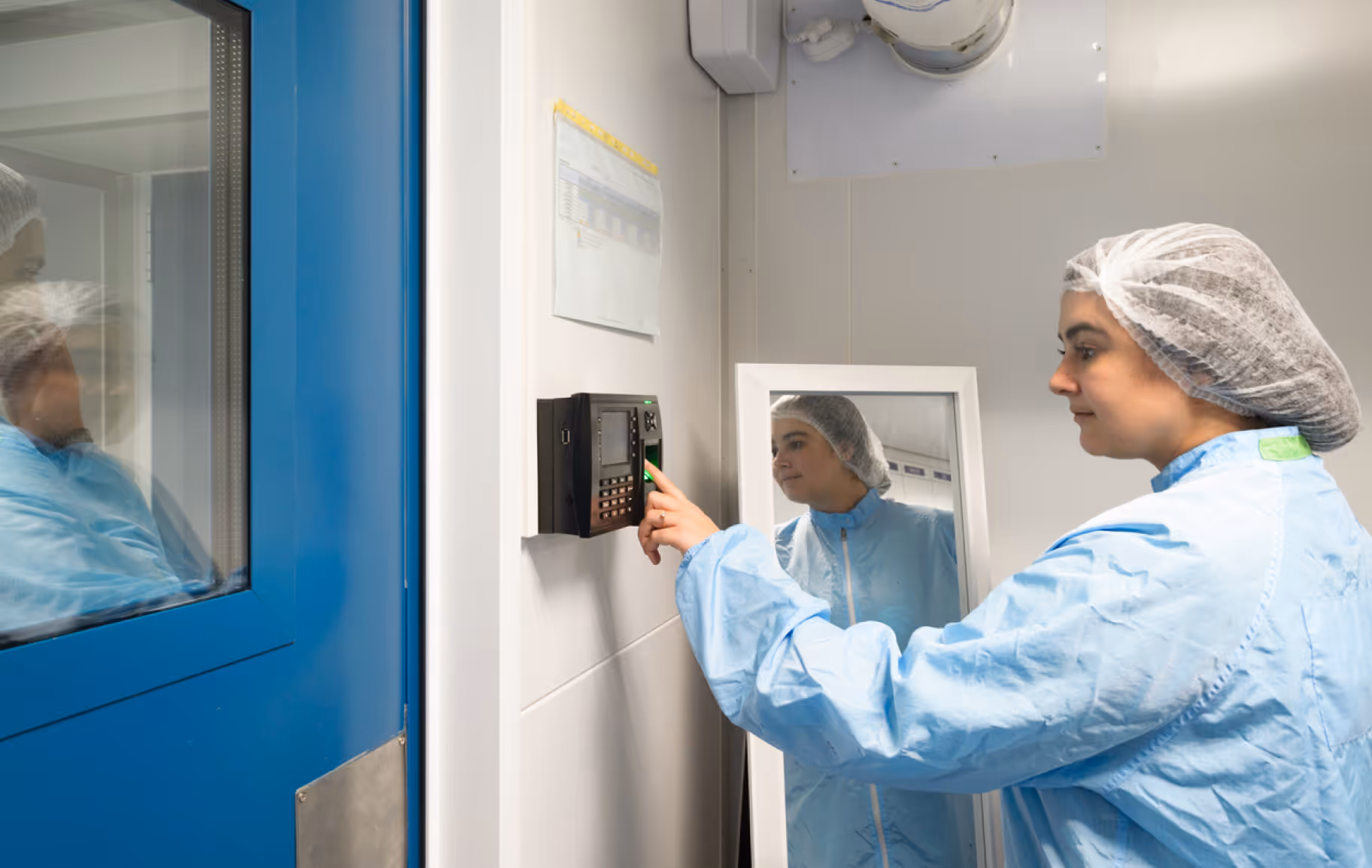 Healthcare worker in blue protective clothing and hair net using a fingerprint scanner on a wall next to a blue door.