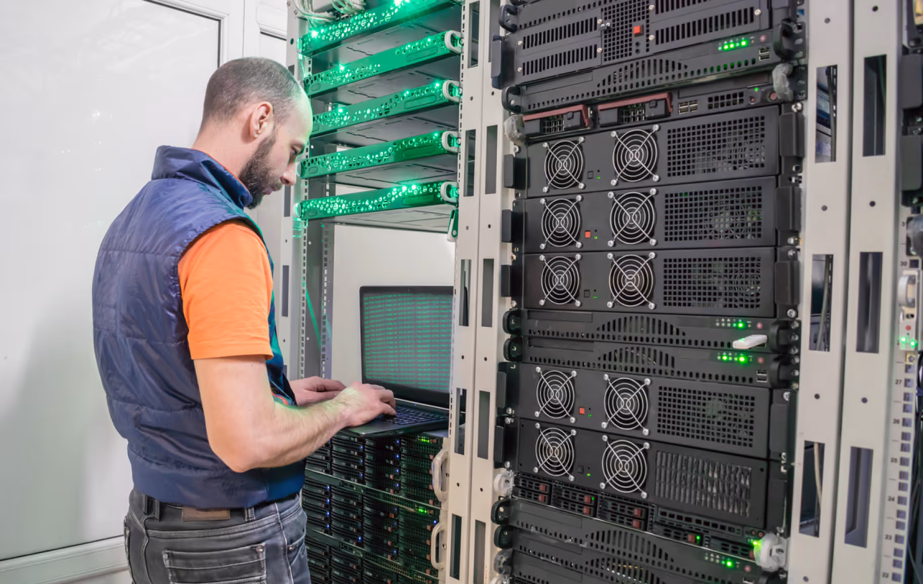 Technician working on a laptop inside a server room with racks of servers and green indicator lights.