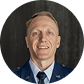 Smiling man wearing a blue uniform jacket with a white shirt and tie against a dark background.