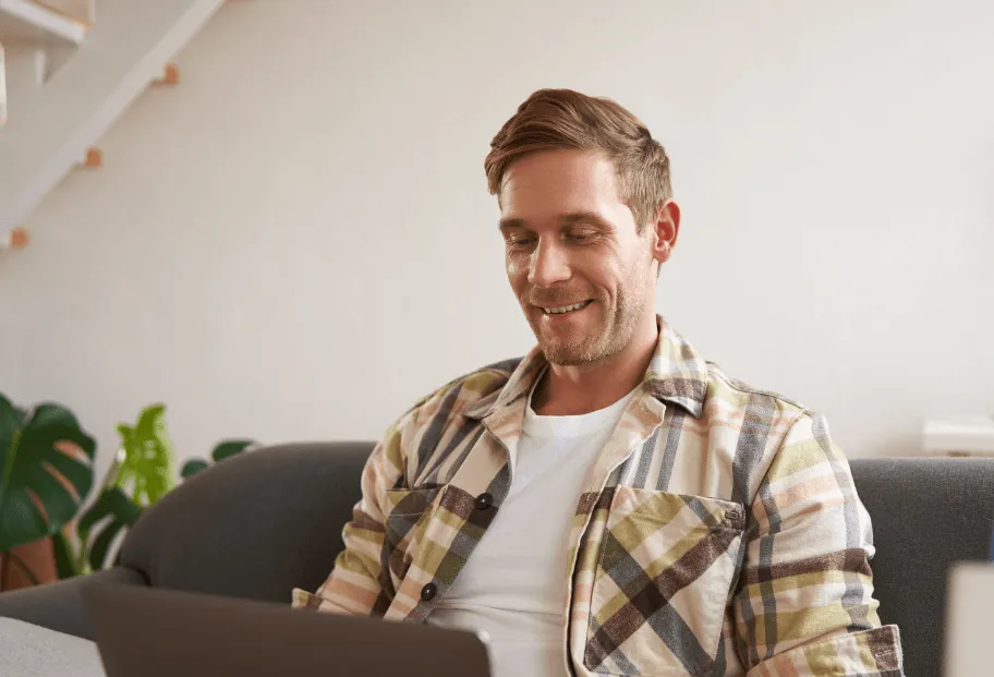 Smiling man sitting on a couch using a laptop in a cozy living room.