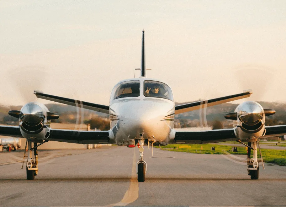 Small twin-engine airplane on runway with propellers spinning and pilot making a peace sign in the cockpit.