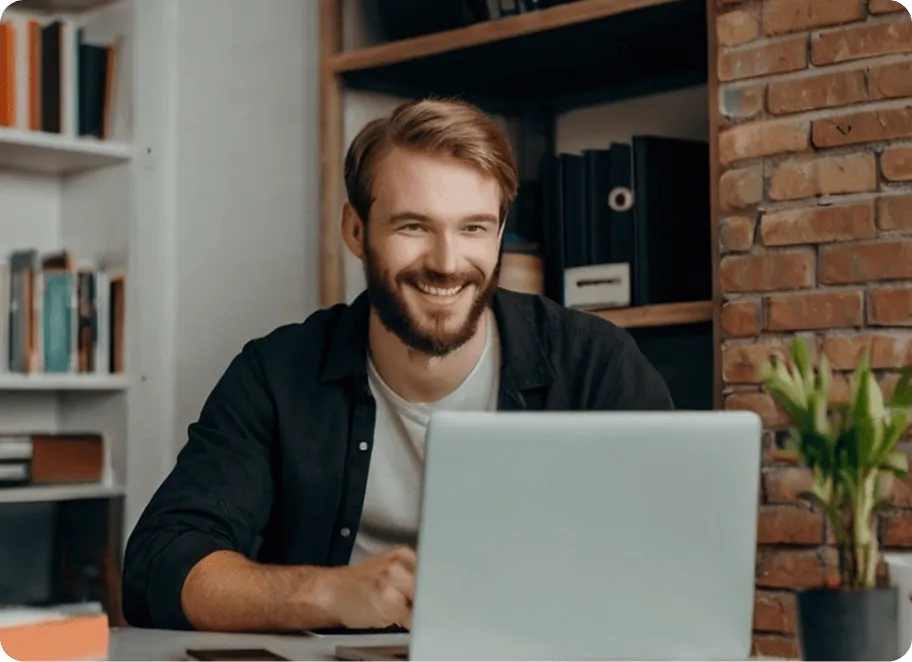 Smiling man with beard working on a laptop at a desk in a cozy room with bookshelves and a plant.