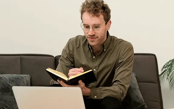 Man with glasses sitting on a couch reading a notebook while looking at a laptop screen.