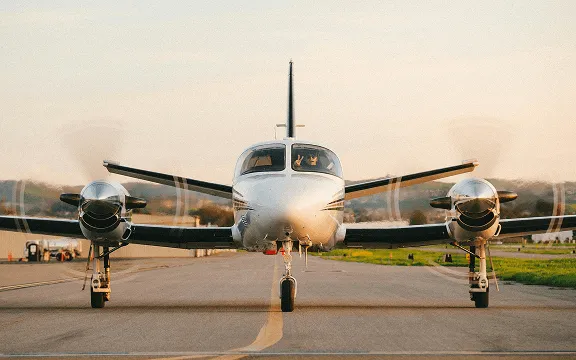 Front view of a twin-engine propeller airplane on a runway at sunset with spinning propellers.