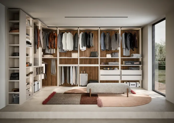 Modern walk-in closet with white shelves and racks holding jackets, shirts, pants, a suitcase, and accessories, next to a bench and large window.