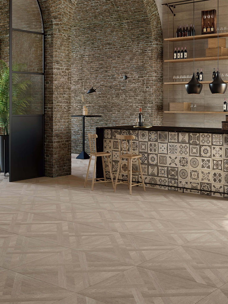 Modern bar area with patterned tile front, two wooden stools, exposed brick walls, hanging black pendant lights, and shelves with wine bottles and glasses.