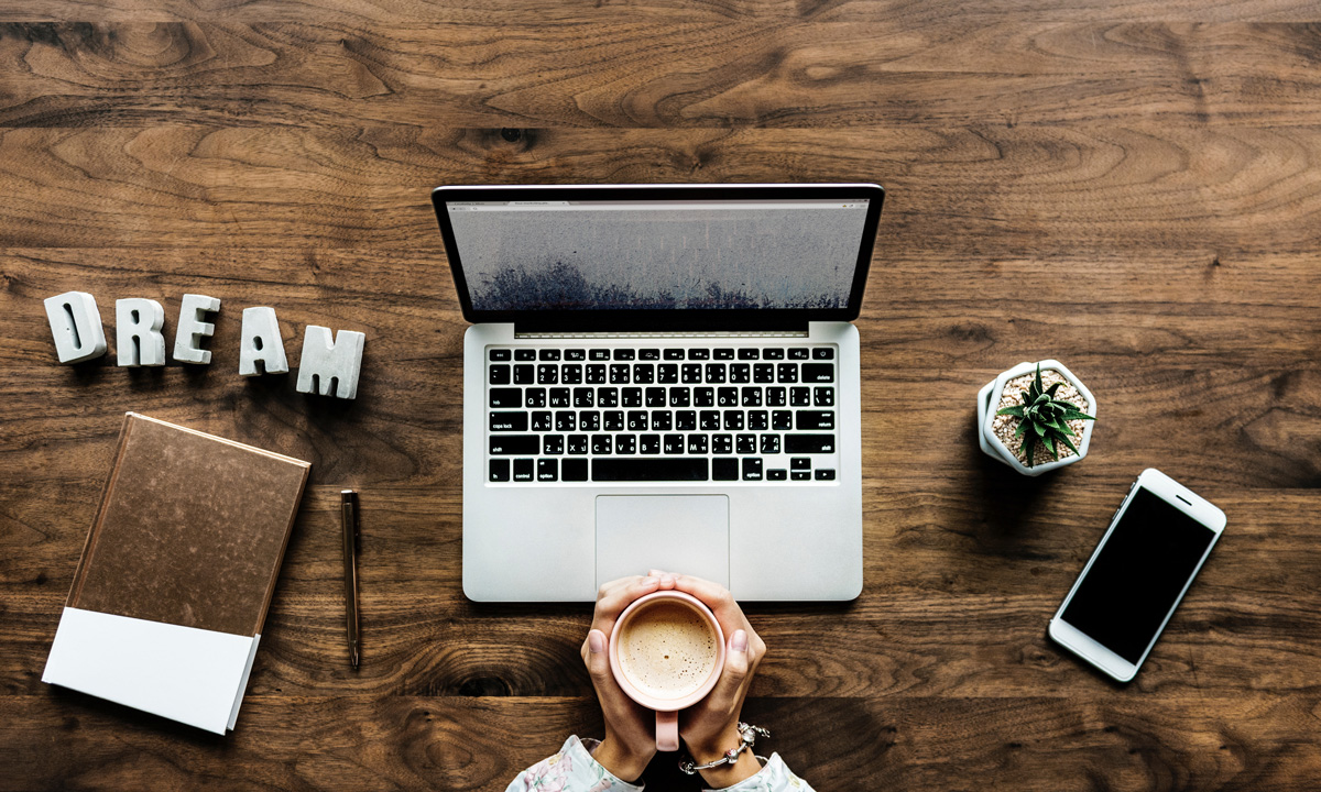 Overhead view of a wooden desk with a laptop, a notebook, a pen, a smartphone, a small potted plant, and hands holding a cup of coffee, with the word DREAM spelled out in block letters.