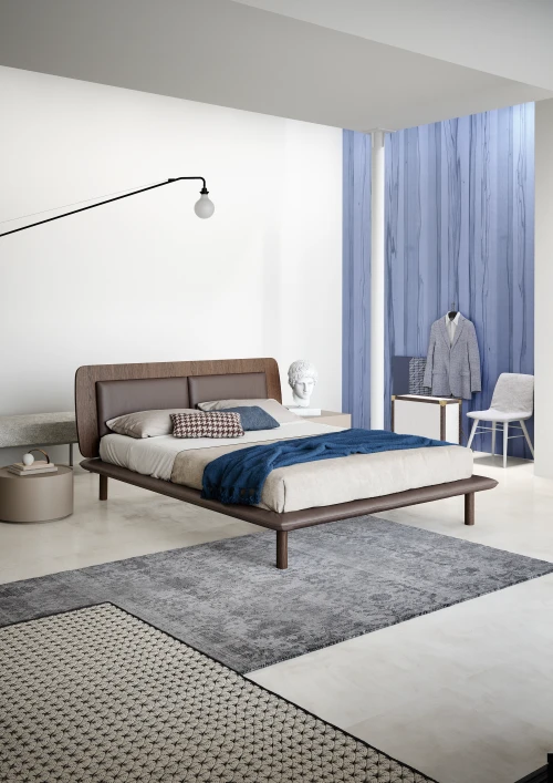Modern bedroom with a low wooden bed featuring beige and brown bedding, a blue throw, two rugs, a minimalist floor lamp, and a blue accent wall with a jacket on a mannequin and a white chair.